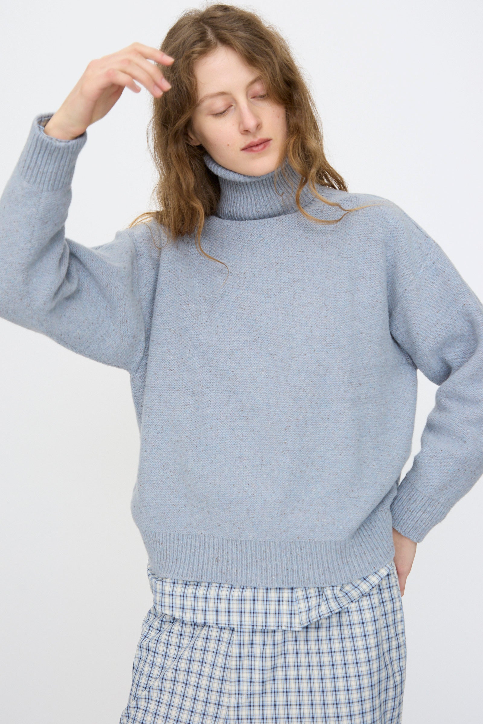 A person in the Baserange Recycled Wool Mea Turtleneck in Ocean and a blue plaid skirt stands with one hand raised and eyes closed, the cozy turtleneck adding warmth to the minimalist background.