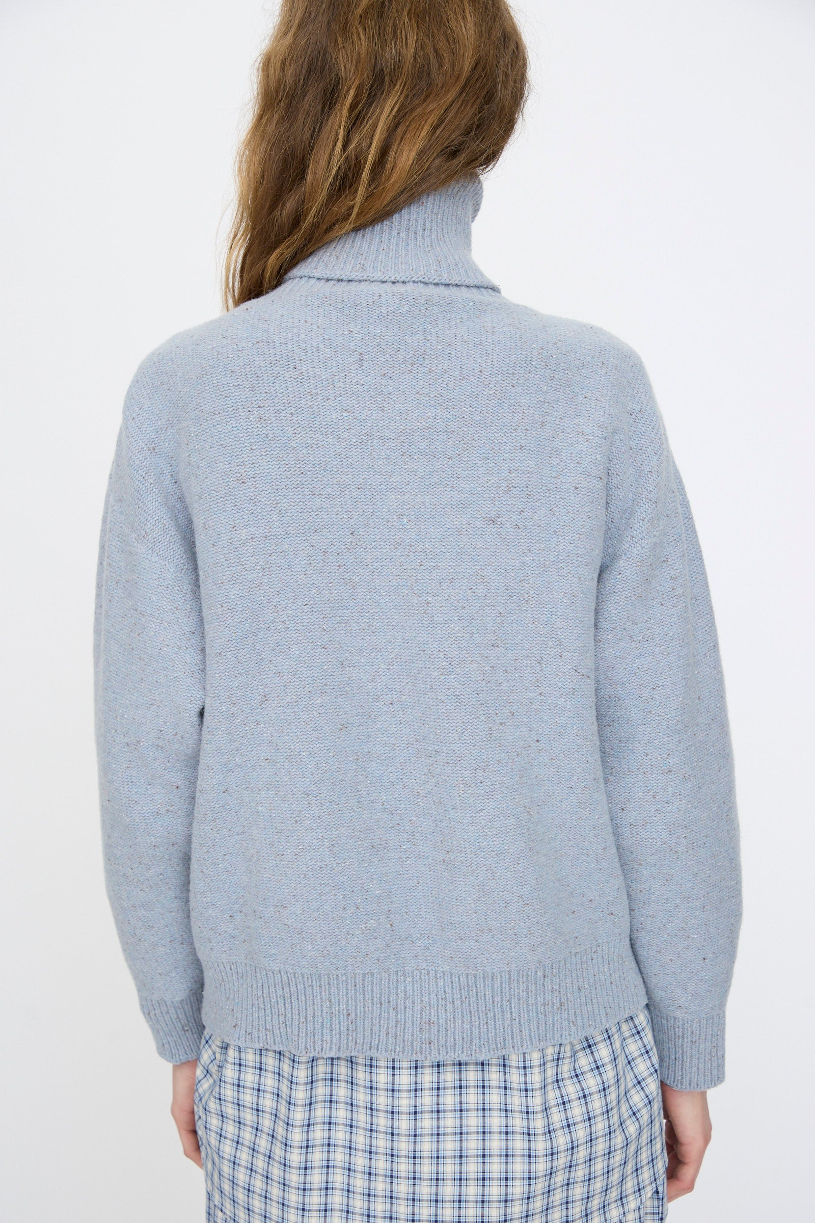 A person with long brown hair, seen from behind, wears the Baserange Recycled Wool Mea Turtleneck in Ocean and a blue and white plaid skirt.