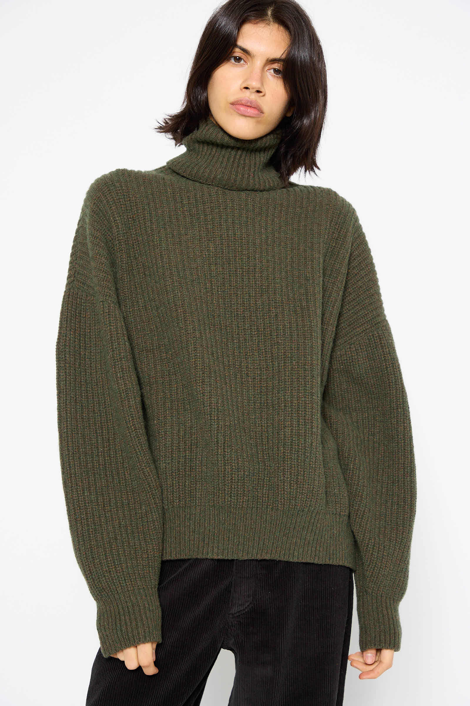 A person wearing the Recycled Wool Mea Turtleneck in Foret by Baserange, a dark green chunky knit sweater made from recycled wool, stands against a plain white background.