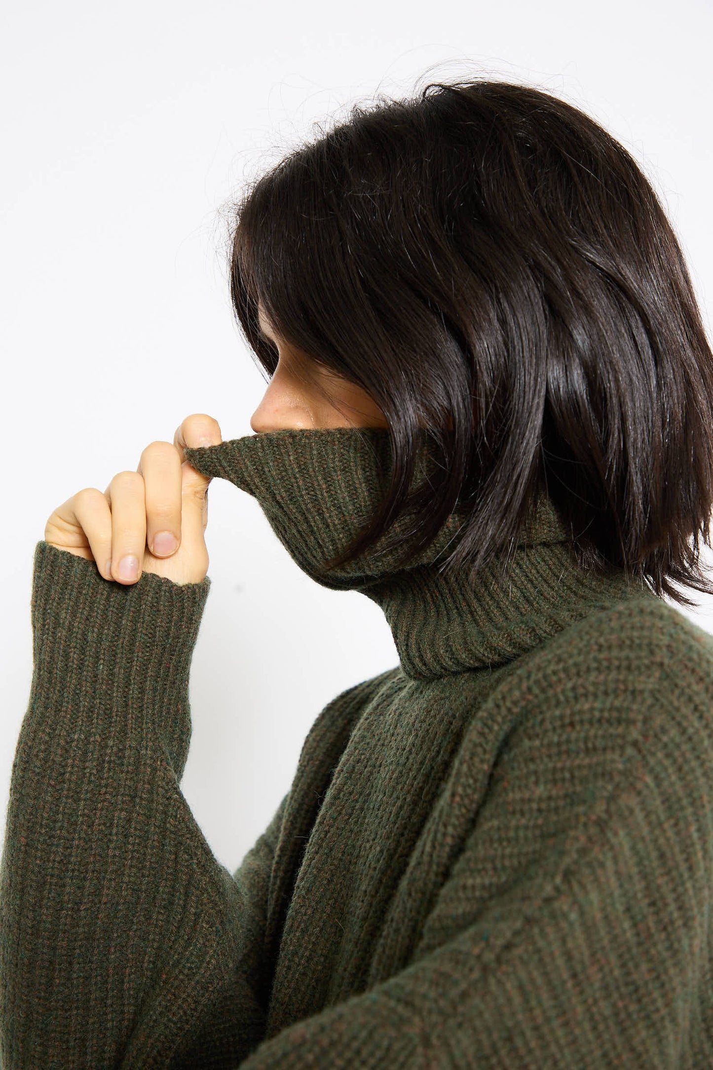 A person with shoulder-length dark hair is seen in profile, partially covering their face with the high collar of the Baserange Recycled Wool Mea Turtleneck in Foret.