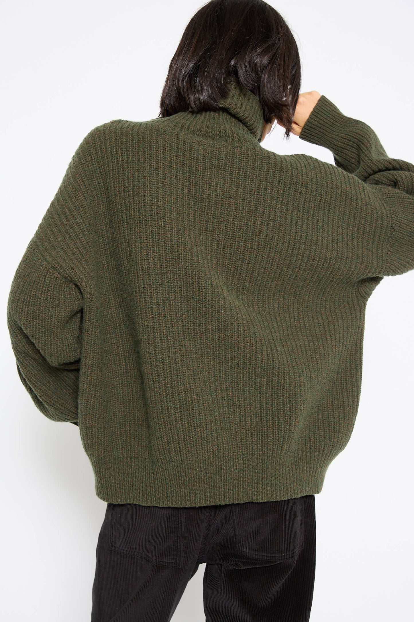 A person with dark hair is facing away from the camera, wearing a Baserange Recycled Wool Mea Turtleneck in Foret and black pants.