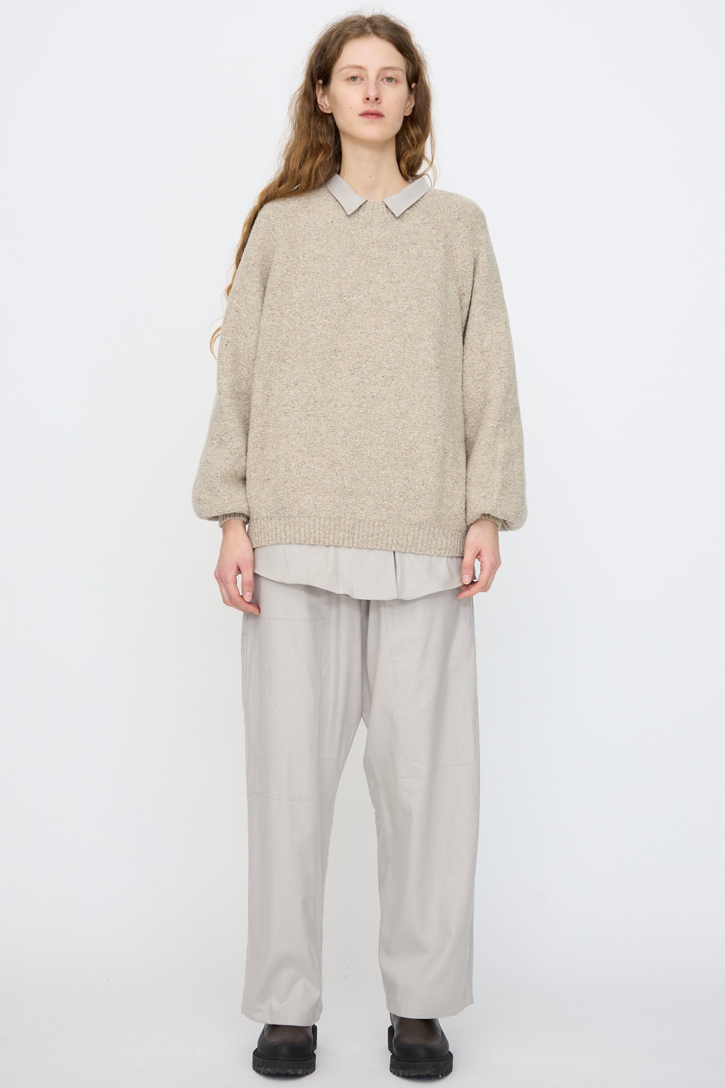 A person stands against a plain background wearing the Baserange Recycled Wool Soar Sweater in Ingra Sand over a collared shirt, with loose light gray pants and black shoes.