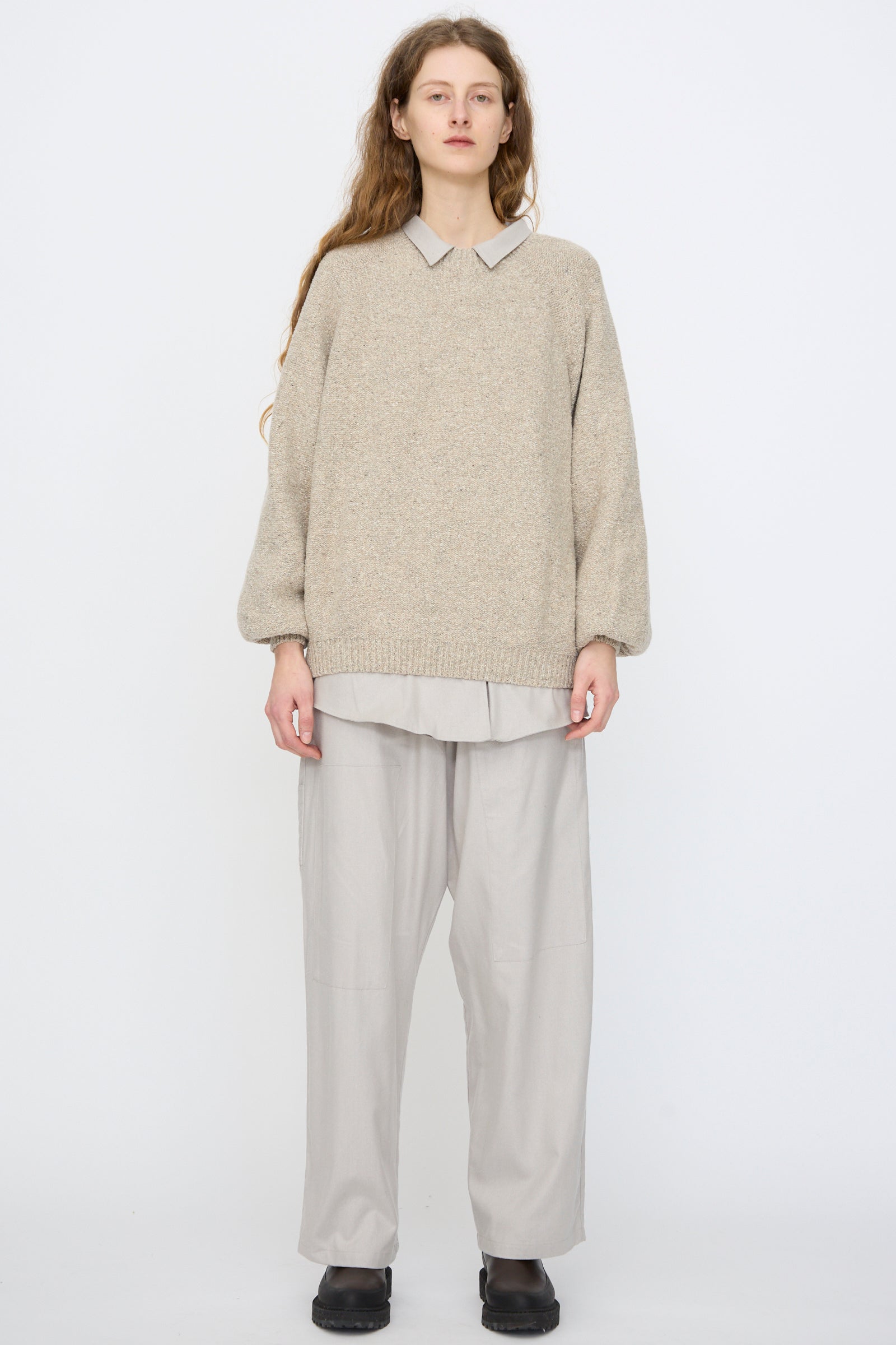 A person stands against a plain background wearing the Baserange Recycled Wool Soar Sweater in Ingra Sand over a collared shirt, with loose light gray pants and black shoes.
