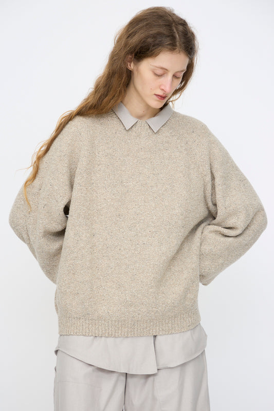 A person with long brown hair looks down, wearing Baserange’s Recycled Wool Soar Sweater in Ingra Sand over a light gray collared shirt and matching loose pants, standing against a plain white background.