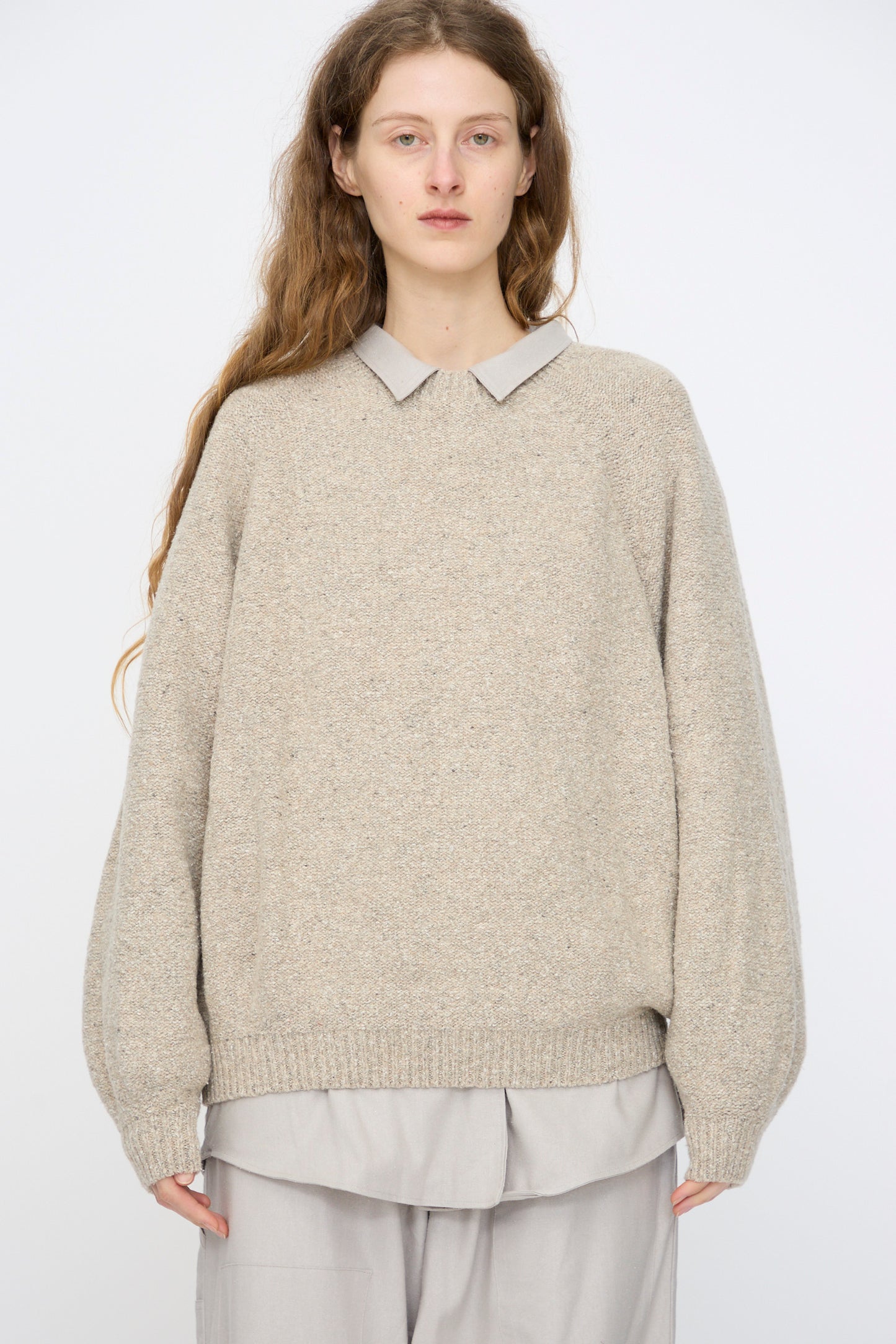 A person with long brown hair wears the Baserange Recycled Wool Soar Sweater in Ingra Sand, styled over a light collared shirt and wide-leg pants, standing against a plain white background.
