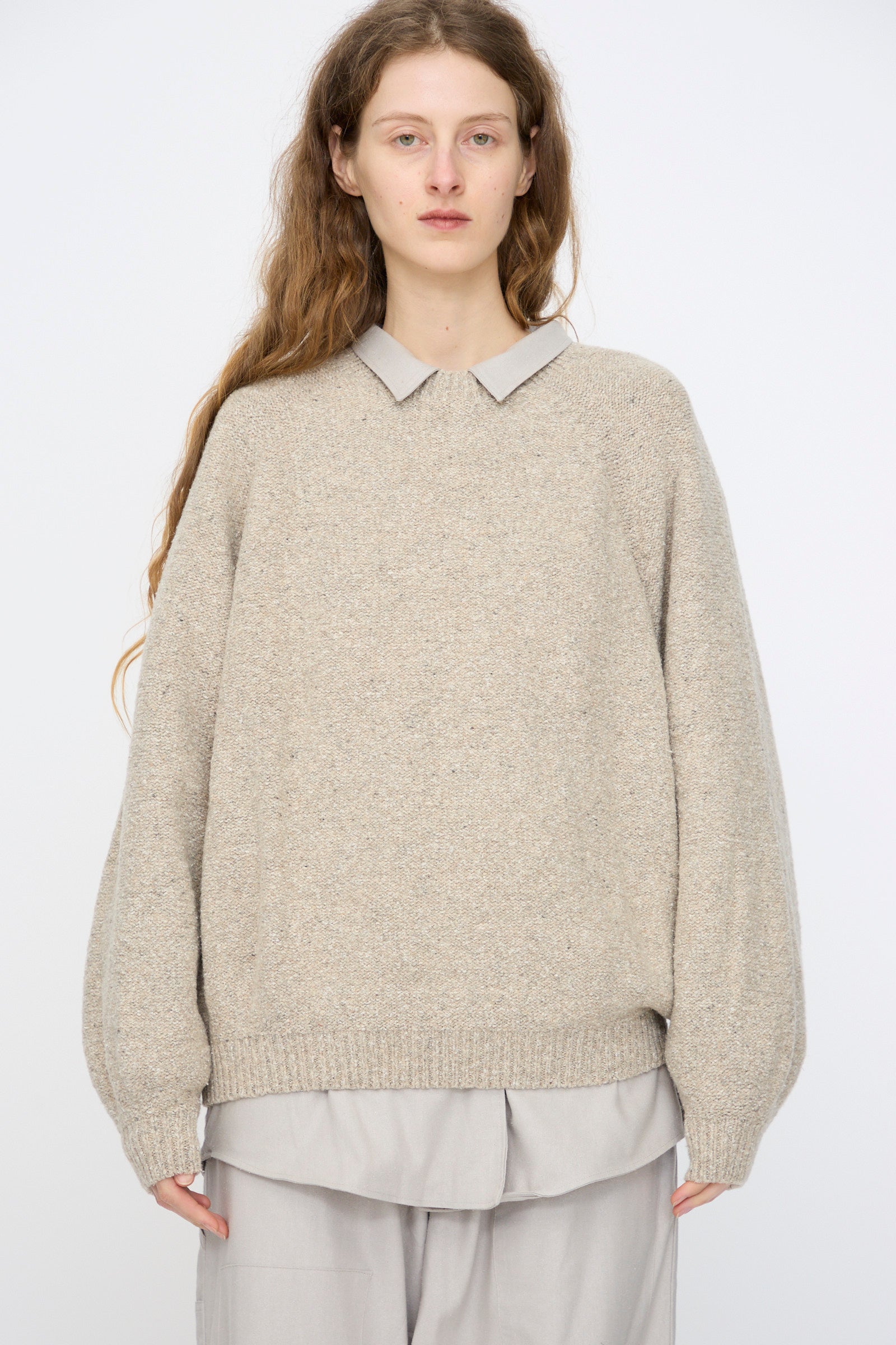 A person with long brown hair wears the Baserange Recycled Wool Soar Sweater in Ingra Sand, styled over a light collared shirt and wide-leg pants, standing against a plain white background.