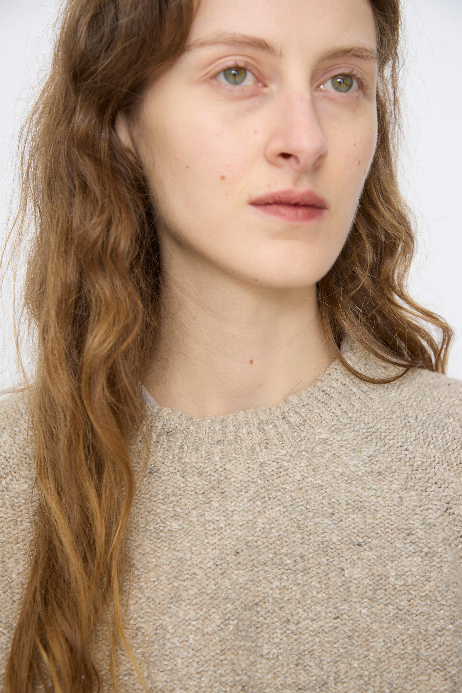 A person with long, wavy brown hair wears the Baserange Recycled Wool Soar Sweater in Ingra Sand, made in France, gazing slightly to the side against a plain background.