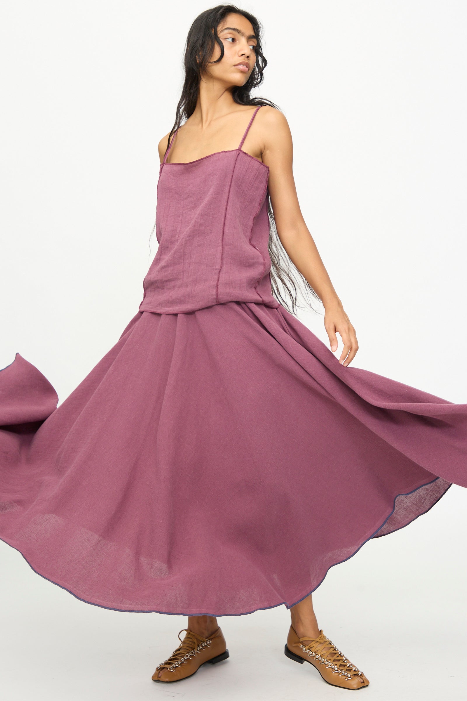A woman stands against a plain background, looking to the side and holding out the skirt of the Baserange Rene Skirt in Aubergine, styled with brown woven shoes.