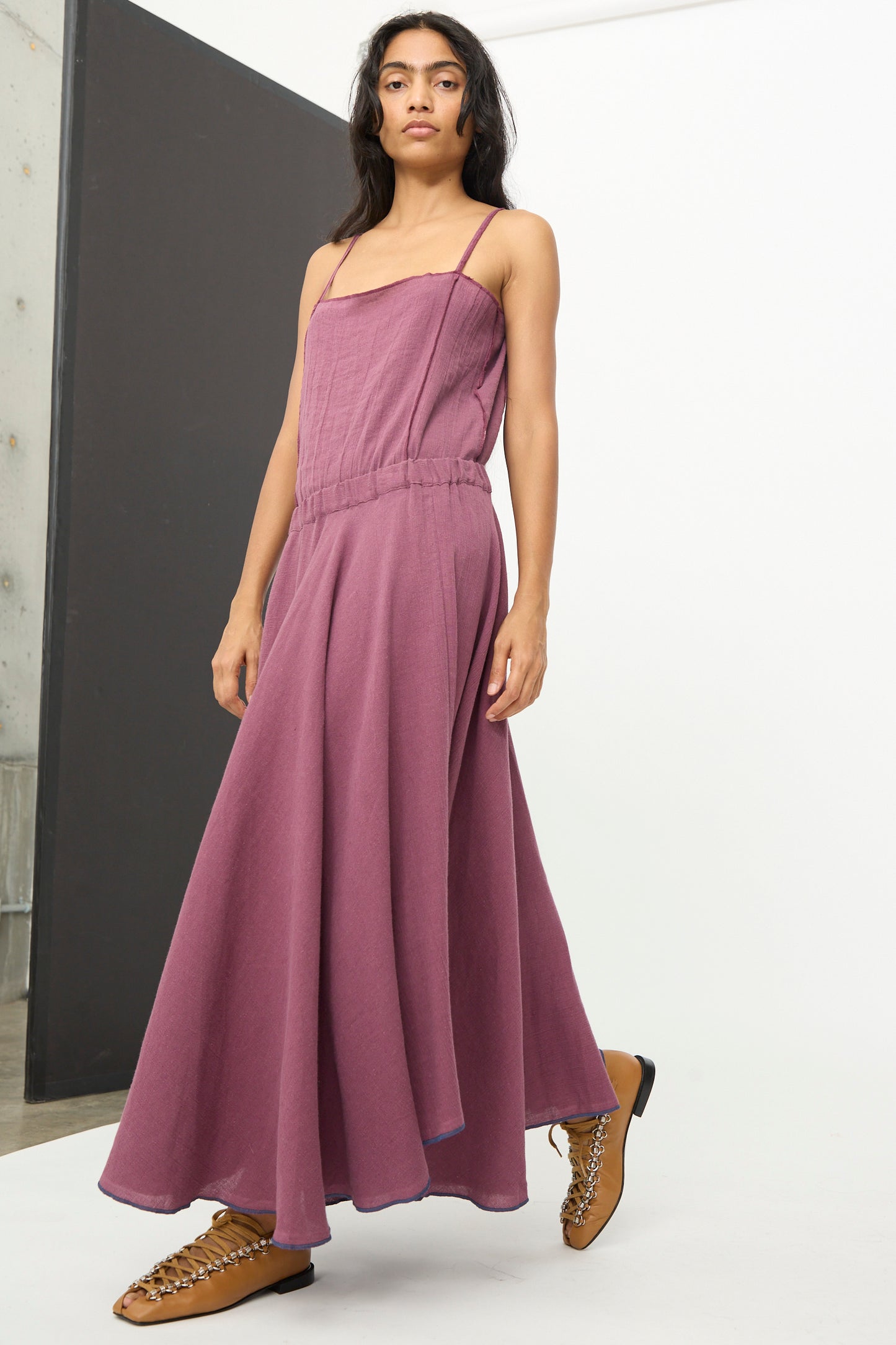A person stands indoors against a plain background wearing the Baserange Rene Skirt in Aubergine, styled with tan lace-up shoes.