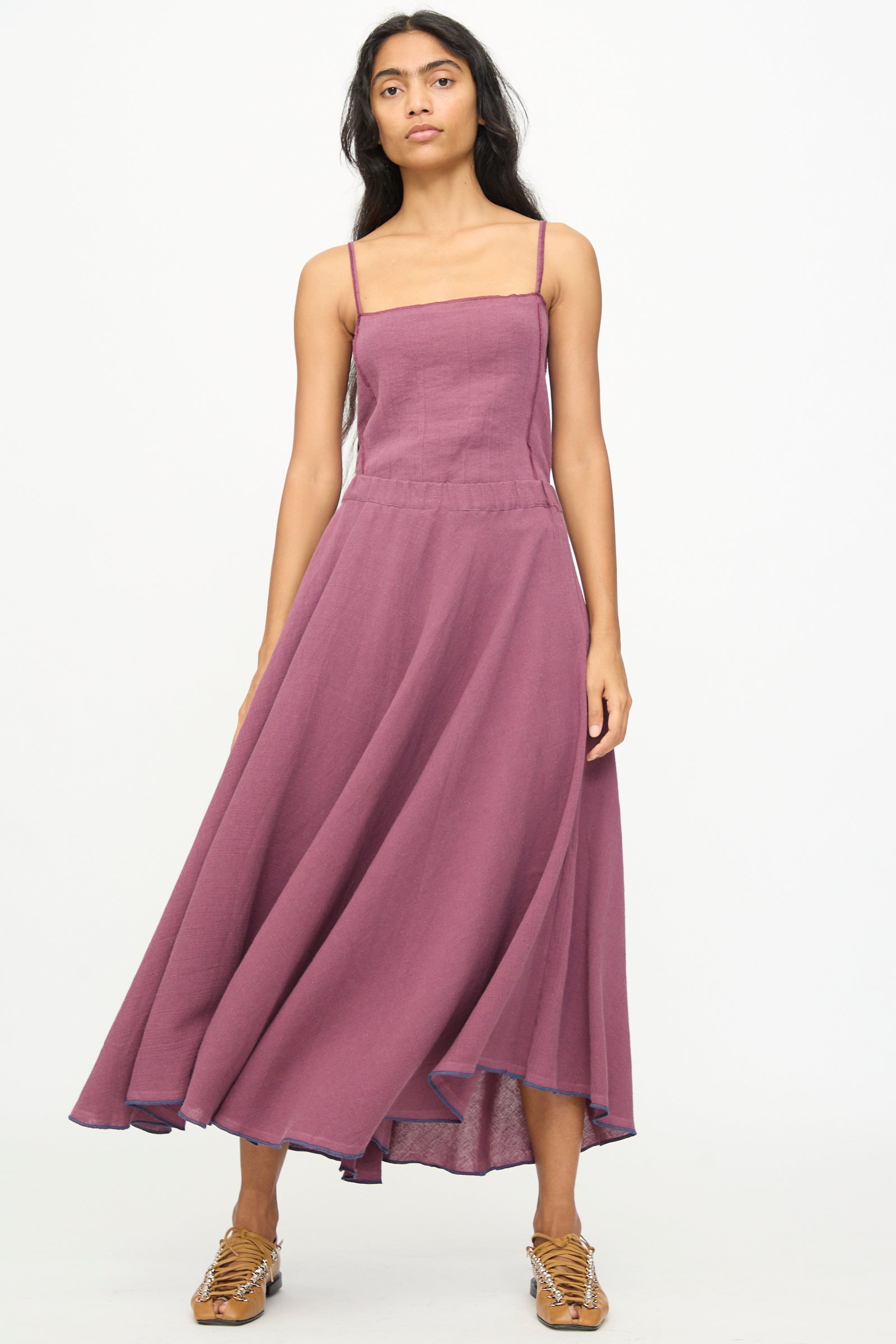 A woman wearing the Baserange Rene Skirt in Aubergine, styled with tan woven sandals, stands against a plain white background.