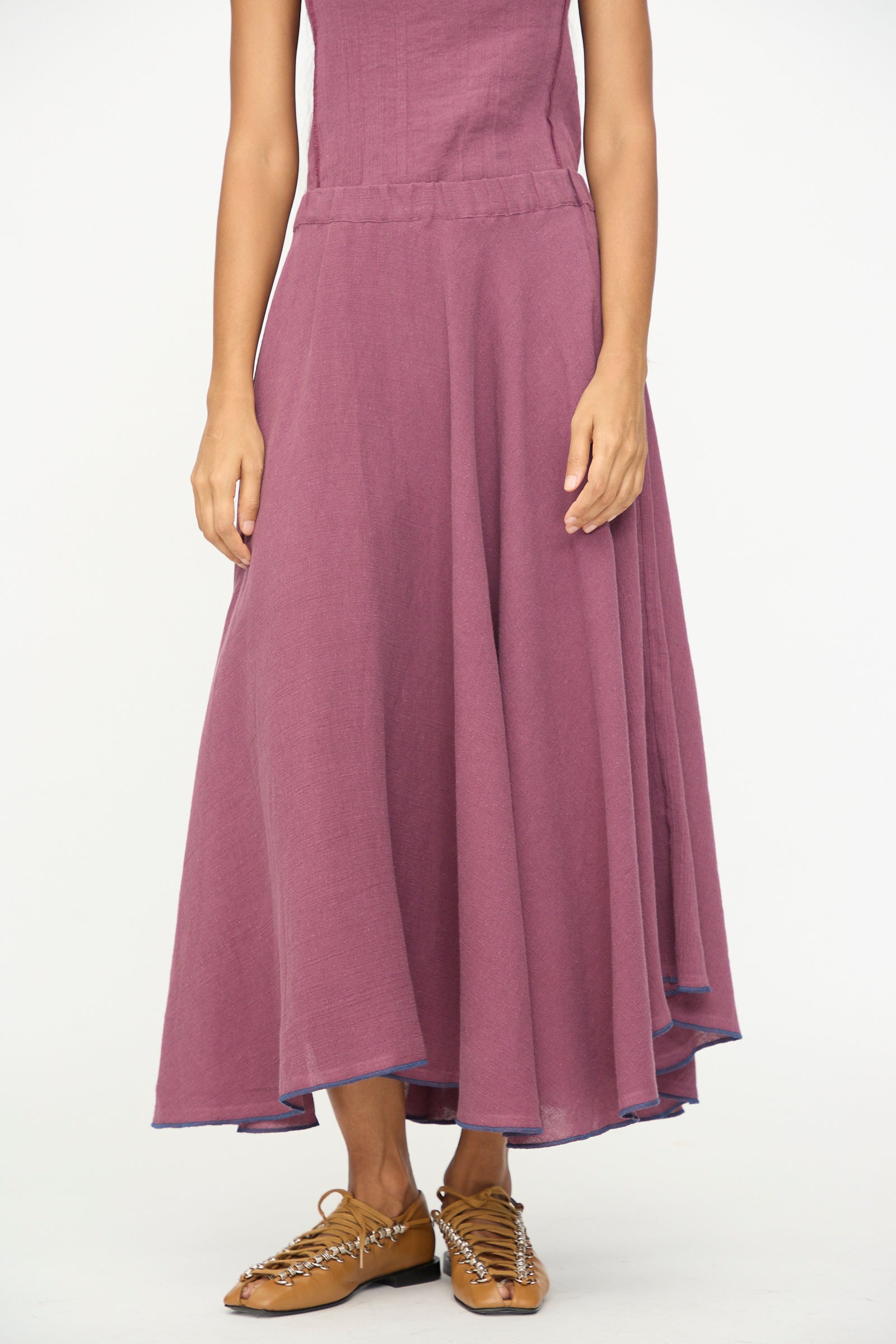 A person wearing the Baserange Rene Skirt in Aubergine, paired with tan woven flat shoes, stands against a plain white background.