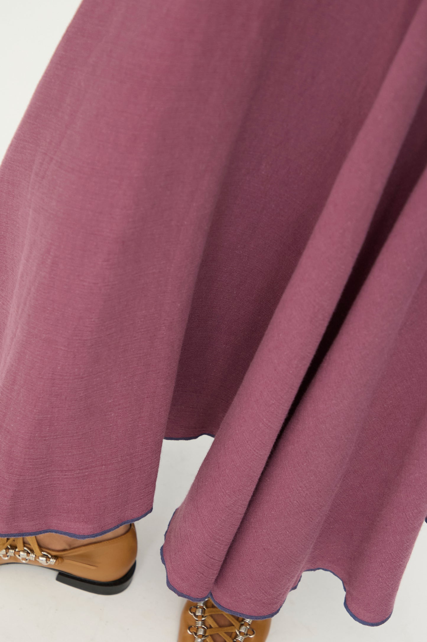 A close-up of the Baserange Rene Skirt in Aubergine, a mauve-colored midi skirt with a slight flare and an asymmetrical hem, crafted from a soft linen-cotton blend, paired with tan lace-up shoes on a light background.
