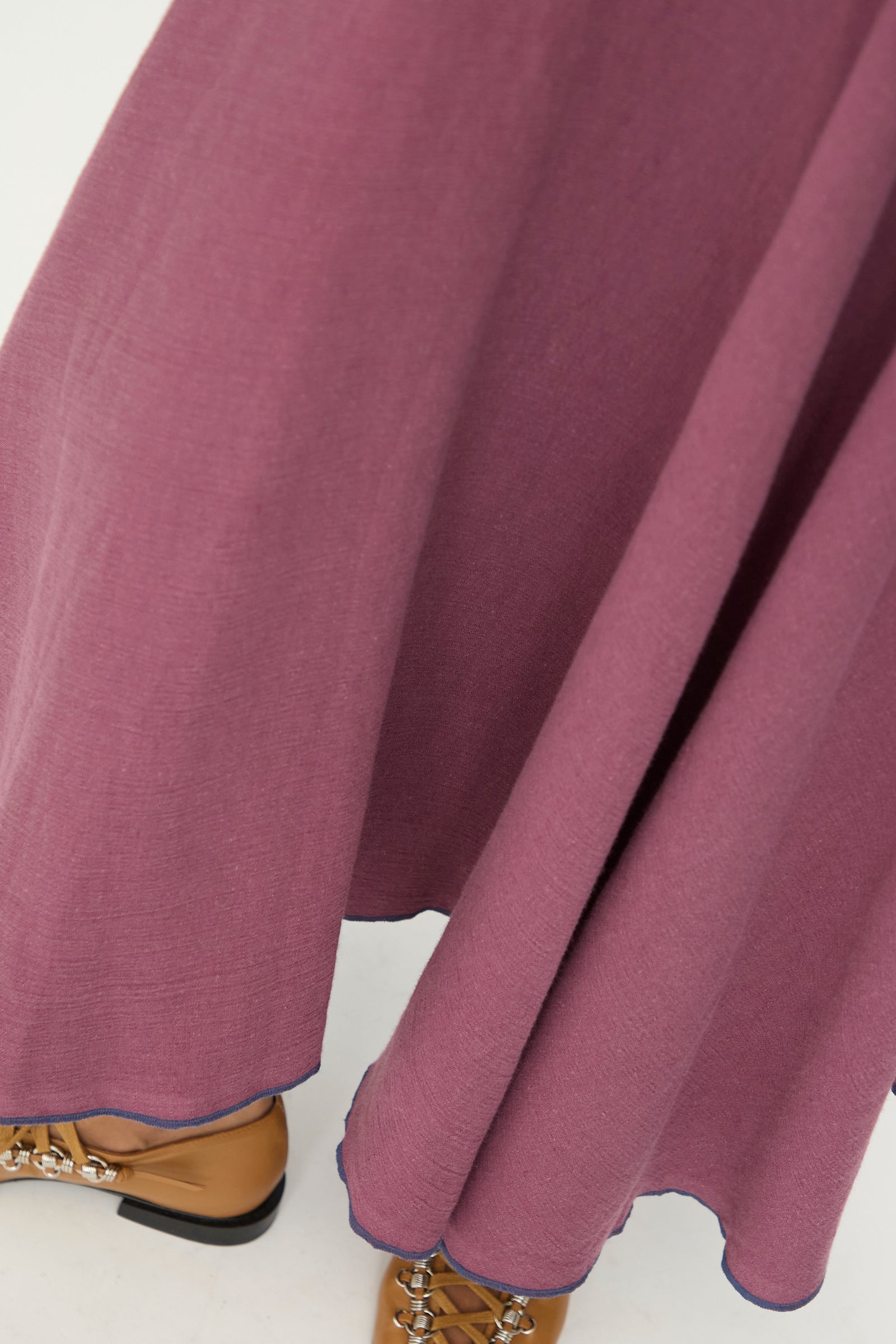A close-up of the Baserange Rene Skirt in Aubergine, a mauve-colored midi skirt with a slight flare and an asymmetrical hem, crafted from a soft linen-cotton blend, paired with tan lace-up shoes on a light background.