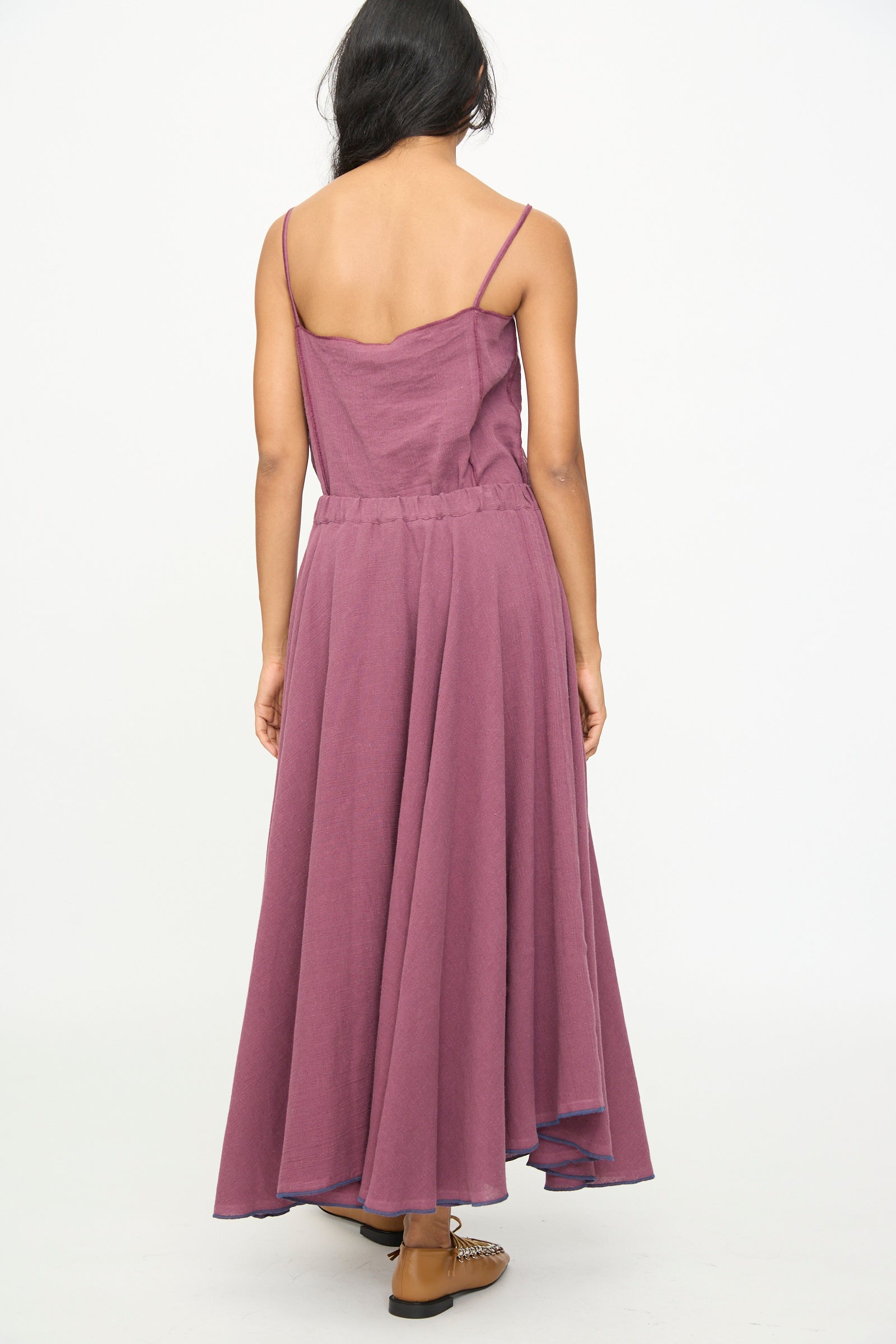 A person stands with their back to the camera, wearing the Baserange Rene Skirt in Aubergine—an ankle-length piece crafted from a linen cotton blend—paired with brown shoes, against a plain white background.