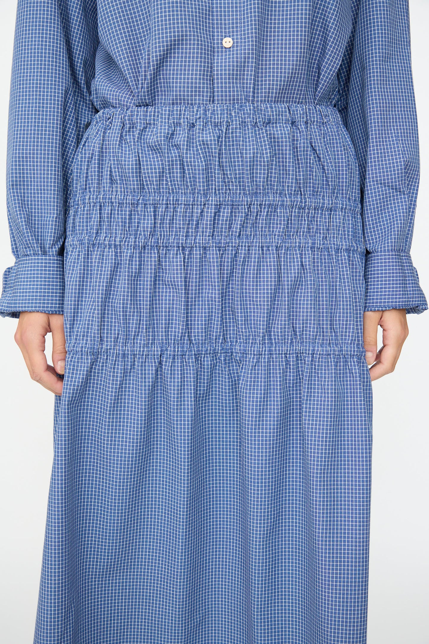 A person wears the Baserange Rezi Skirt in Ace Check, a blue checkered piece crafted from a lyocell cotton blend and featuring gathered, ruched detailing at the waist and skirt. The image is cropped to show the skirt from the chest to the knees.