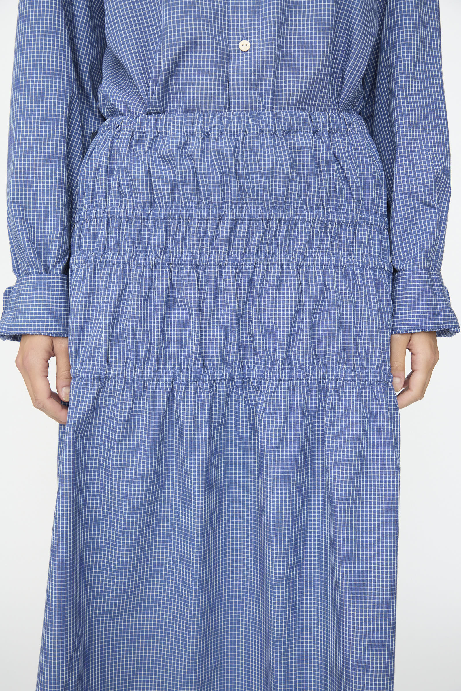 A person wears the Baserange Rezi Skirt in Ace Check, a blue checkered piece crafted from a lyocell cotton blend and featuring gathered, ruched detailing at the waist and skirt. The image is cropped to show the skirt from the chest to the knees.