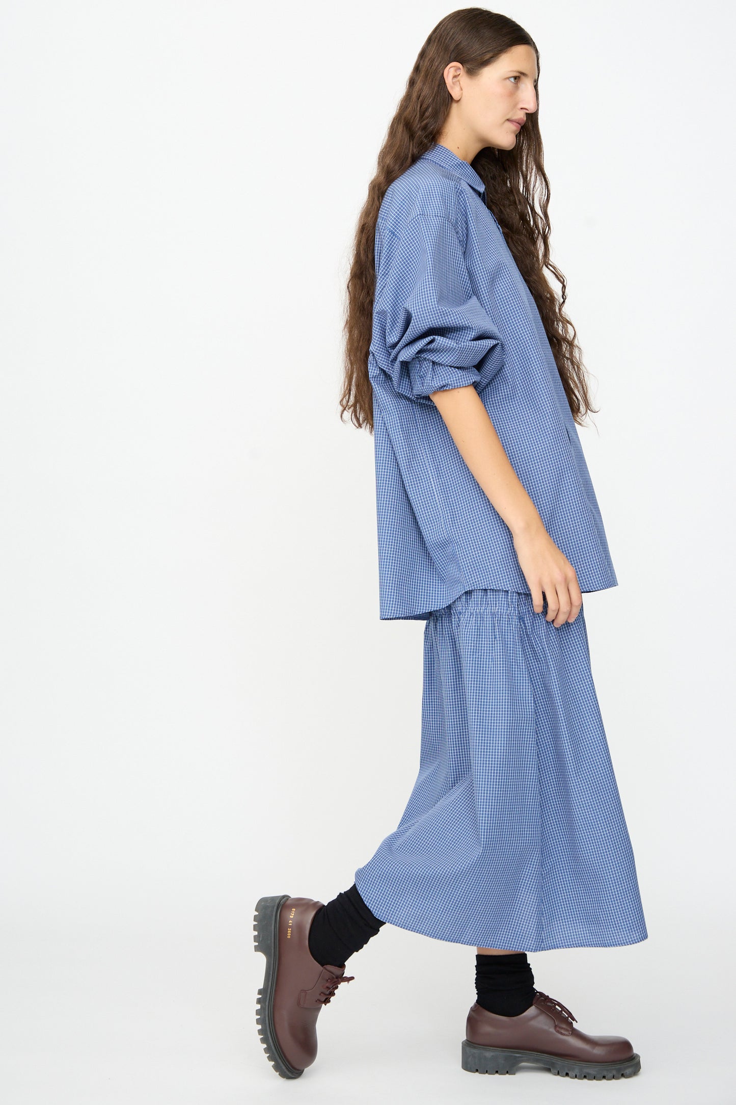 Person with long hair wearing a loose blue striped lyocell cotton blend shirt, the Baserange Rezi Skirt in Ace Check—a matching midi skirt with an elastic waistband—black socks, and brown shoes, walking against a plain white background.