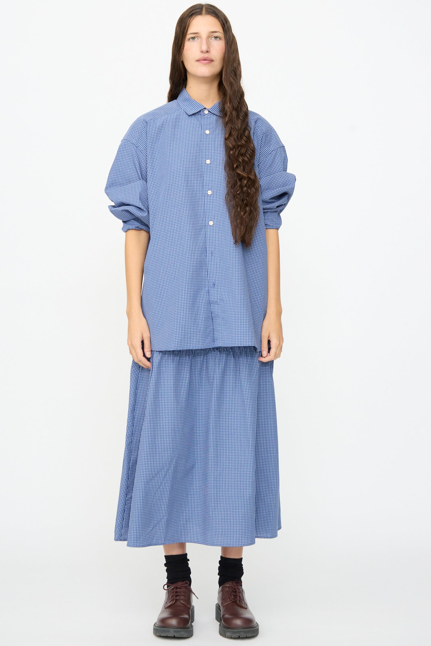 A woman stands against a white background, wearing Baserange’s Rezi Skirt in Ace Check—a blue and white striped button-up midi skirt with an elastic waistband—paired with a matching shirt, brown shoes, and dark socks.