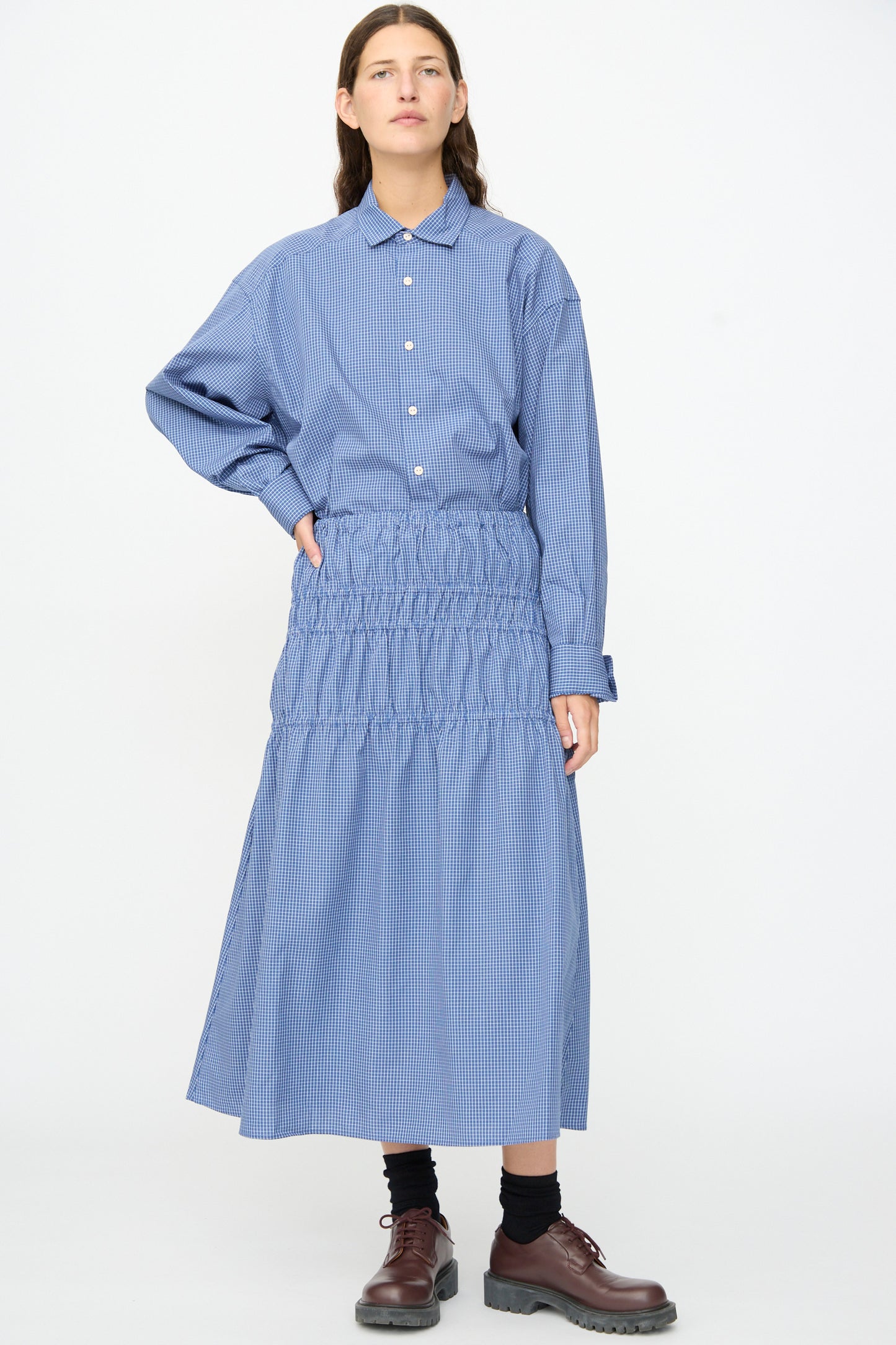 A person wearing a blue long-sleeve button-up shirt and the Baserange Rezi Skirt in Ace Check—a lyocell cotton blend midi skirt with an elastic waistband—along with black socks and brown shoes, stands against a plain white background.
