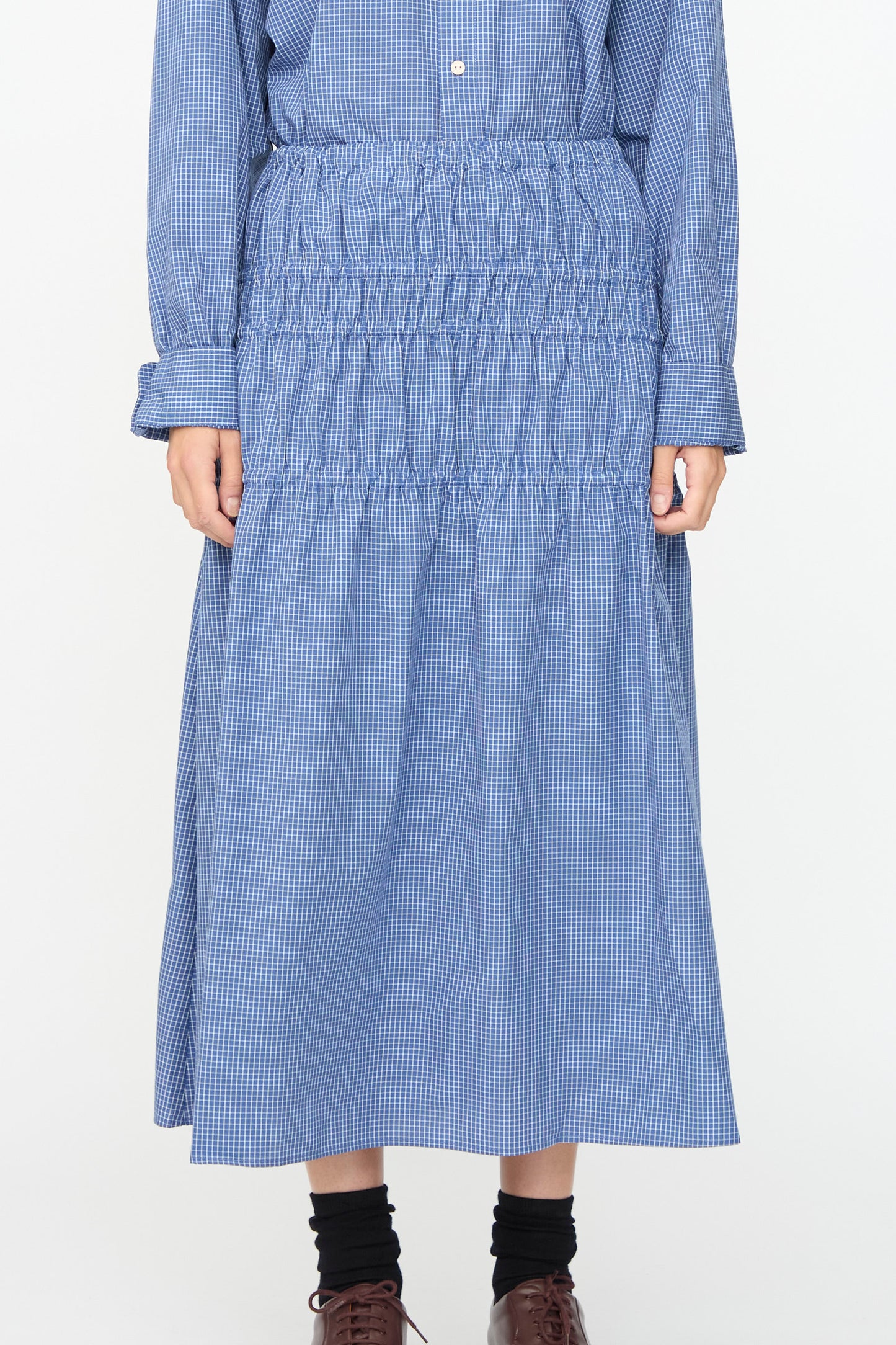 A person wearing a blue gingham long-sleeve shirt and the Baserange Rezi Skirt in Ace Check—a matching smocked midi skirt with an elastic waistband—paired with black socks and brown shoes.