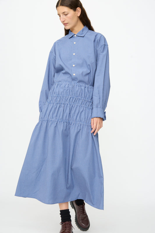 A person wearing the Baserange Rezi Skirt in Ace Check, styled with a collared button-up top and gathered, elastic waistband, paired with dark socks and brown shoes, standing against a plain white background.
