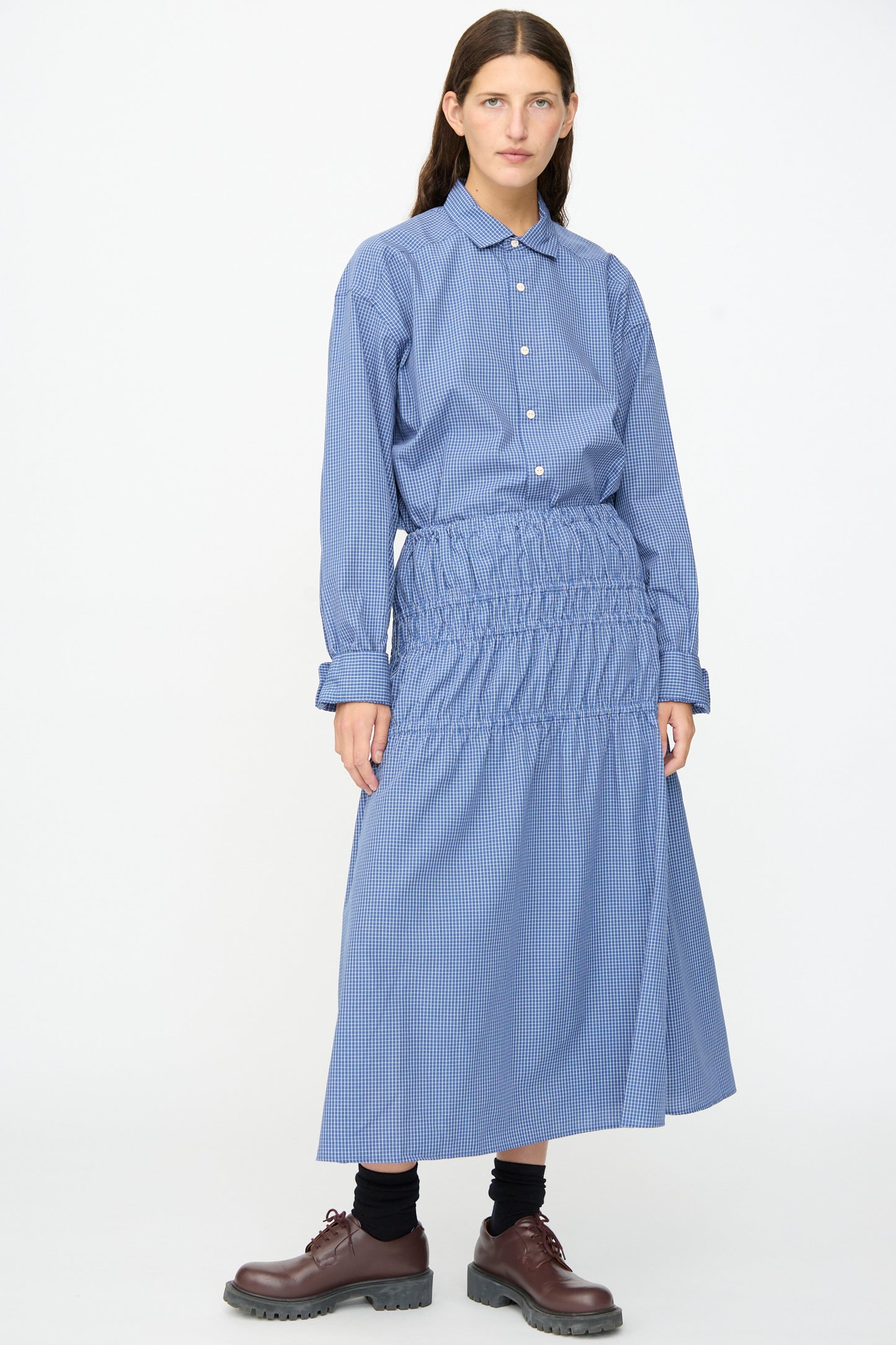 A person wearing a blue, long-sleeve button-up shirt and the Baserange Rezi Skirt in Ace Check—a blue lyocell cotton blend midi skirt with an elastic waistband—paired with black socks and brown shoes, standing against a plain white background.