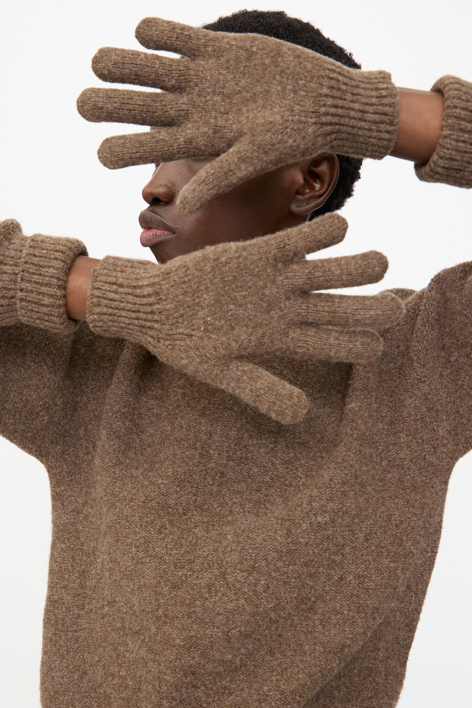 A person wearing Baserange Ribbed Gloves in Burel Brown holds their hands up, partially covering their face against a plain background.