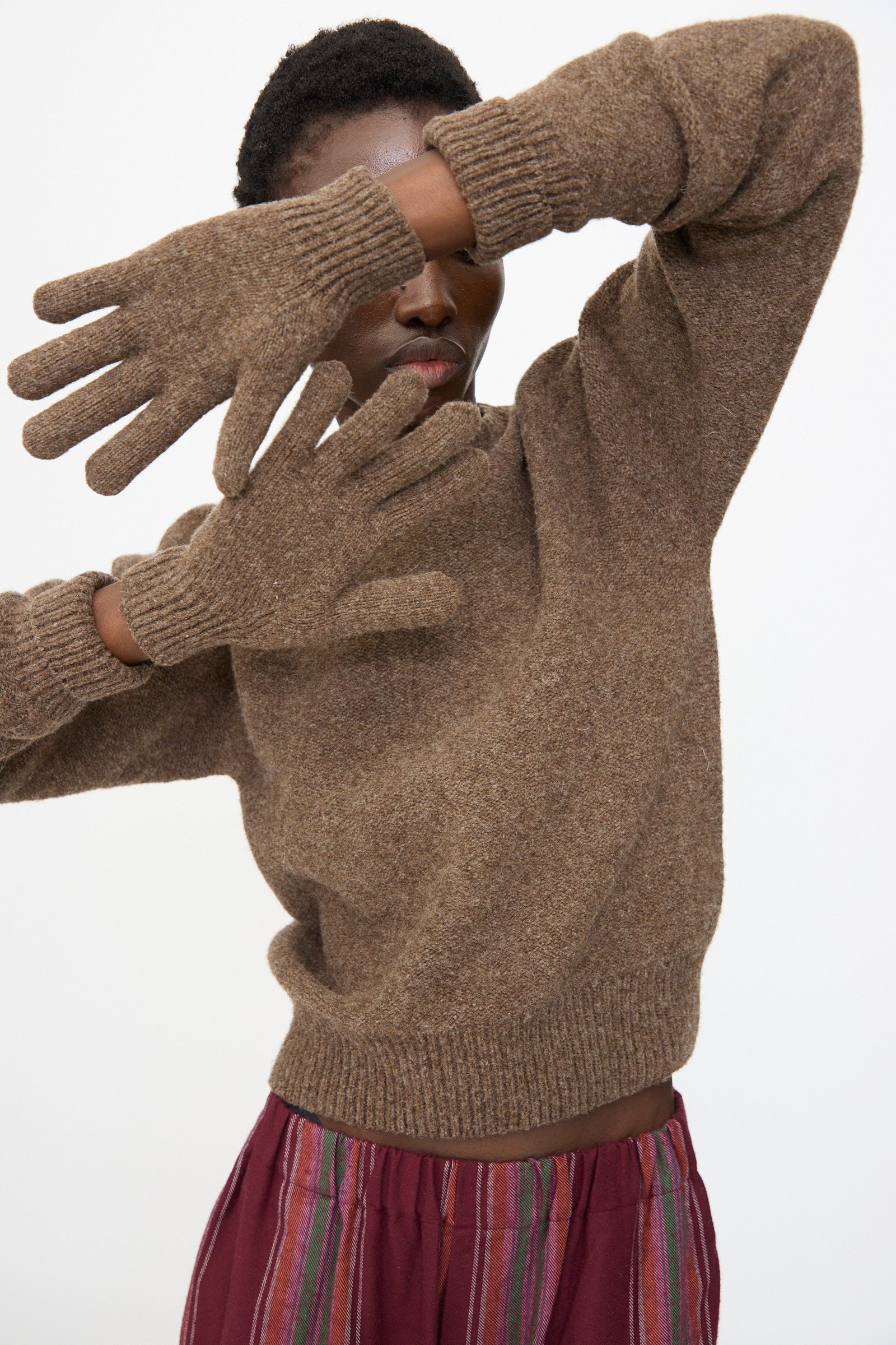 A person wearing Baserange's Ribbed Gloves in Burel Brown poses with arms raised, partially covering their face, and completes the look with a brown knit sweater, burgundy pants, and striped pants.