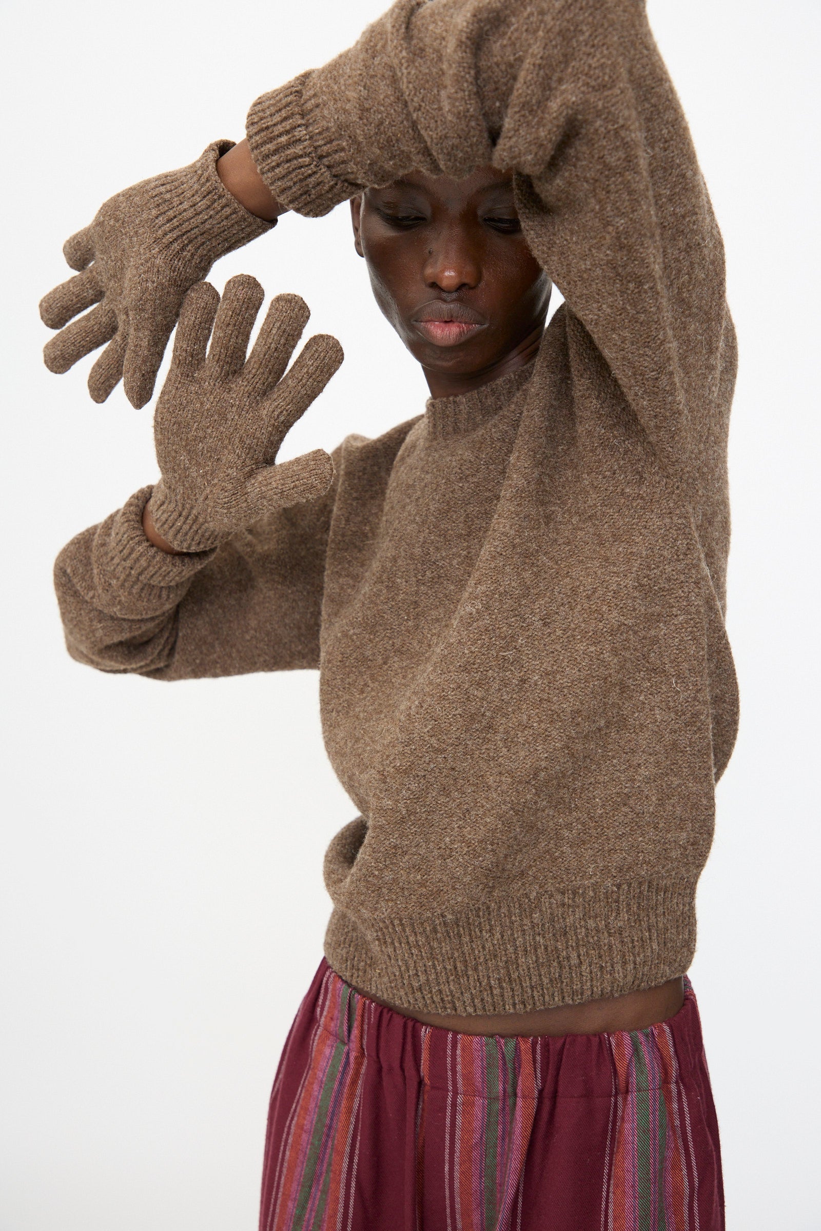 Model wearing Baserange Ribbed Gloves in Burel Brown poses with arms raised, styled with striped maroon pants and a brown sweater against a plain white background.