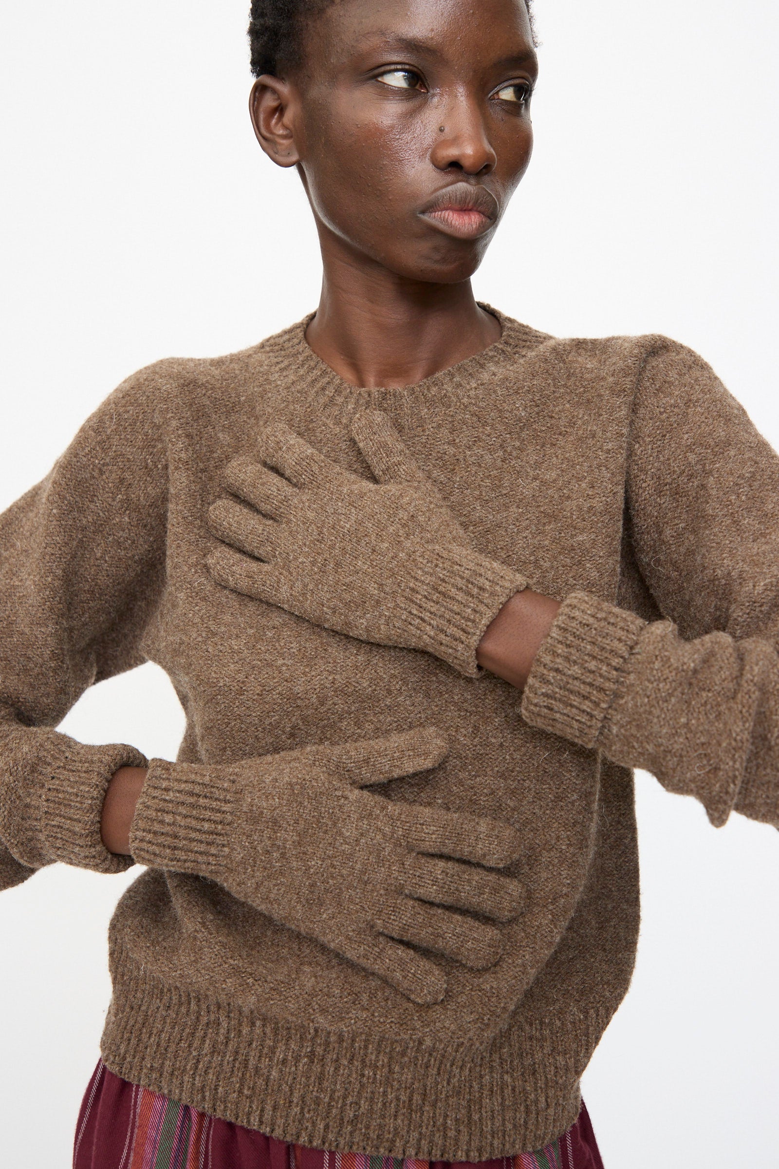 A person wearing Baserange’s Ribbed Gloves in Burel Brown stands with one hand on their chest and the other on their abdomen against a plain background.