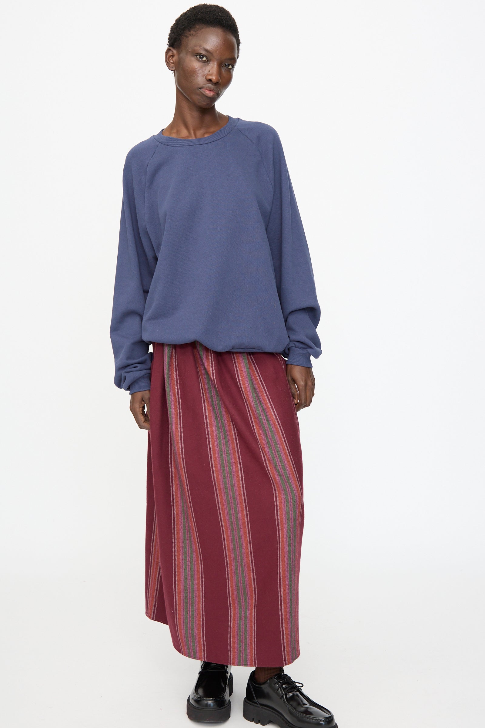 A person stands against a plain background, wearing the Baserange Rim Raglan Sweatshirt in Echo Blue, paired with a long maroon skirt featuring vertical stripes and black shoes.