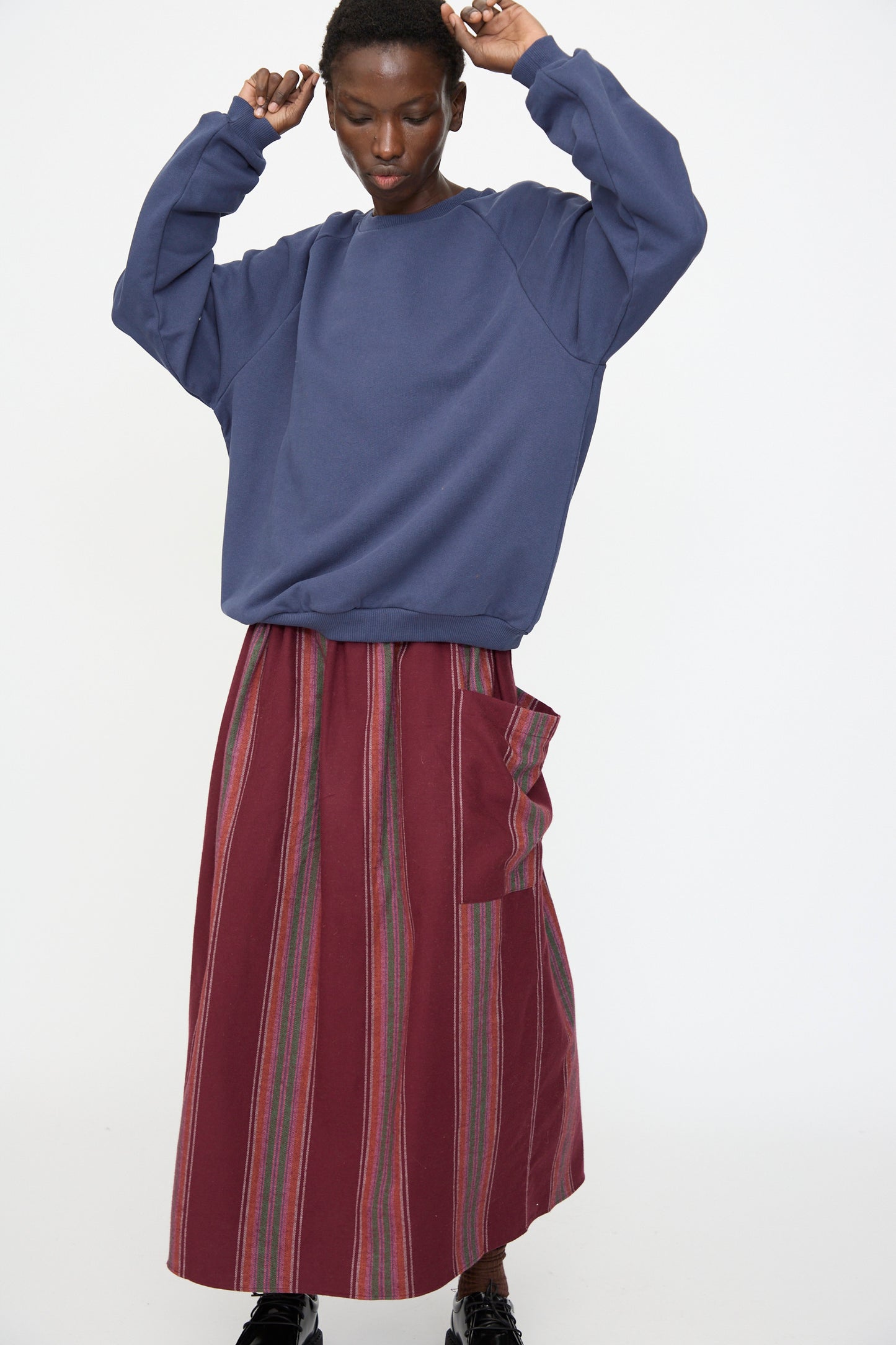 Person wearing the Baserange Rim Raglan Sweatshirt in Echo Blue, styled with a long red skirt featuring vertical stripes and a large side pocket, standing against a plain white background.