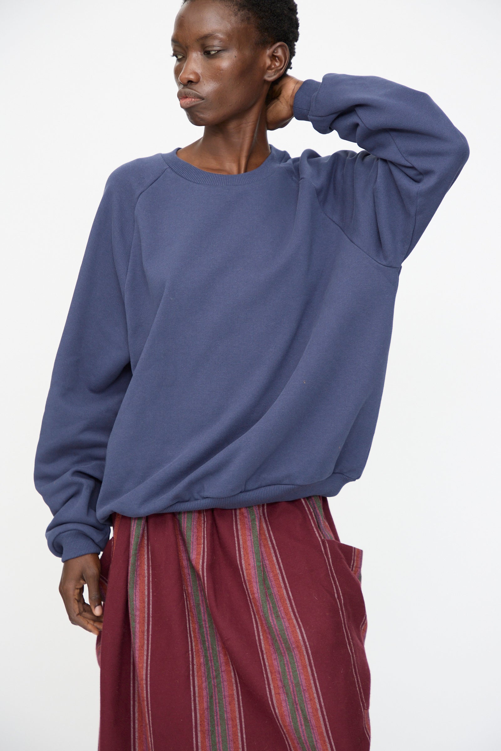 A person wears the Baserange Rim Raglan Sweatshirt in Echo Blue, styled with a maroon skirt featuring vertical multicolored stripes, standing against a plain white background.