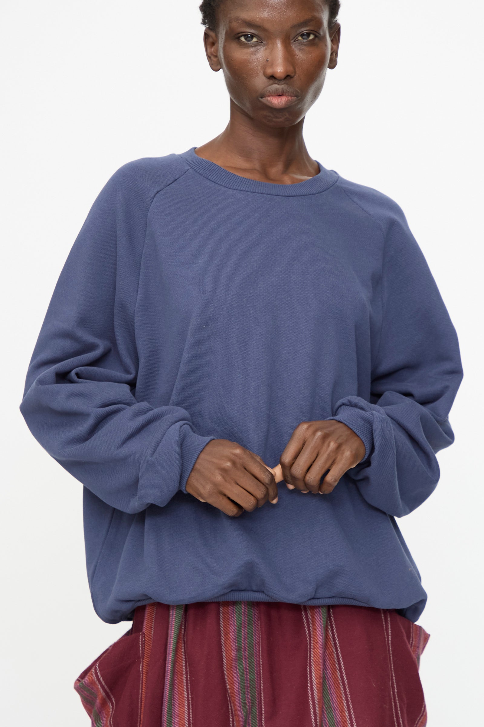A person wearing the Baserange Rim Raglan Sweatshirt in Echo Blue, styled with a red and purple striped skirt, stands against a plain white background.