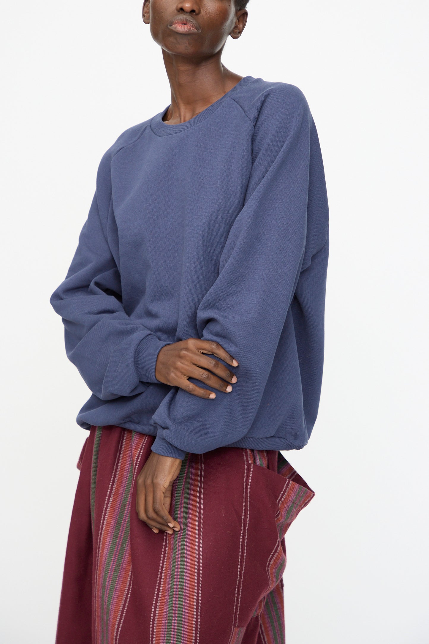 A person wears the Baserange Rim Raglan Sweatshirt in Echo Blue, styled with a maroon vertically striped skirt, standing against a plain white background.