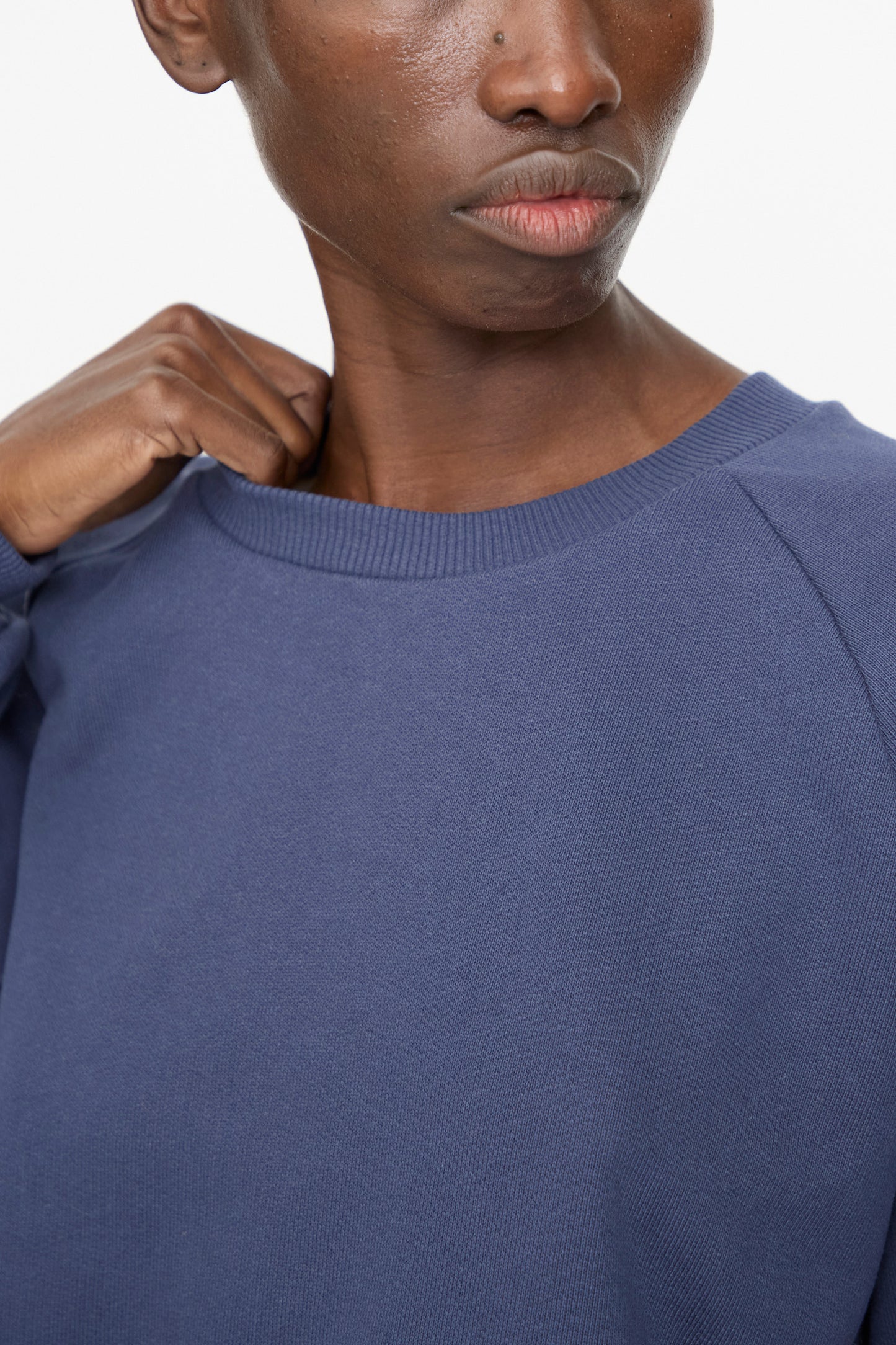 A person wears the Baserange Rim Raglan Sweatshirt in Echo Blue, holding the collar with one hand against a plain white background.