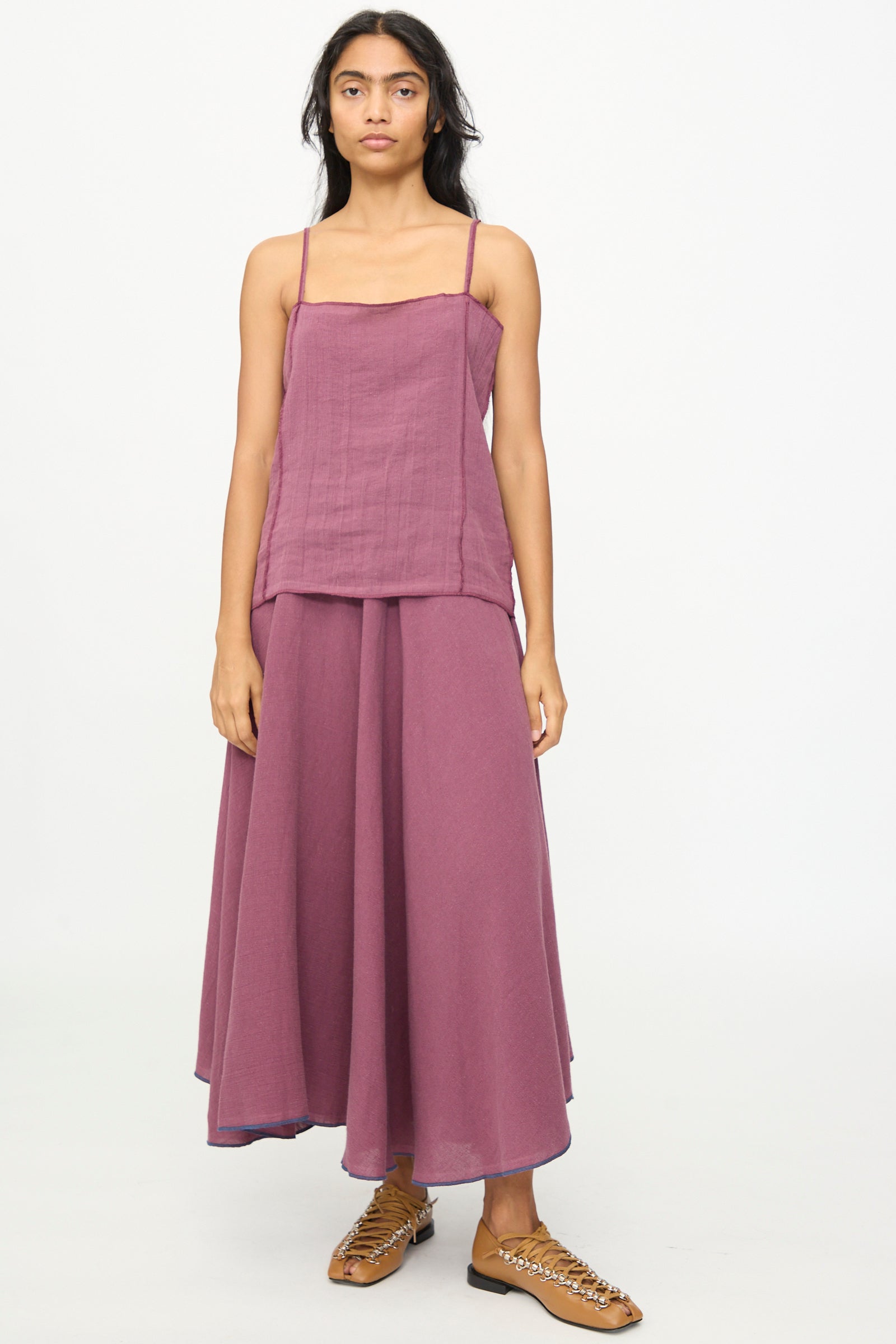 A woman stands facing forward against a plain white background, wearing the Baserange Shok Slip Top in Aubergine with a matching long skirt and tan woven sandals.