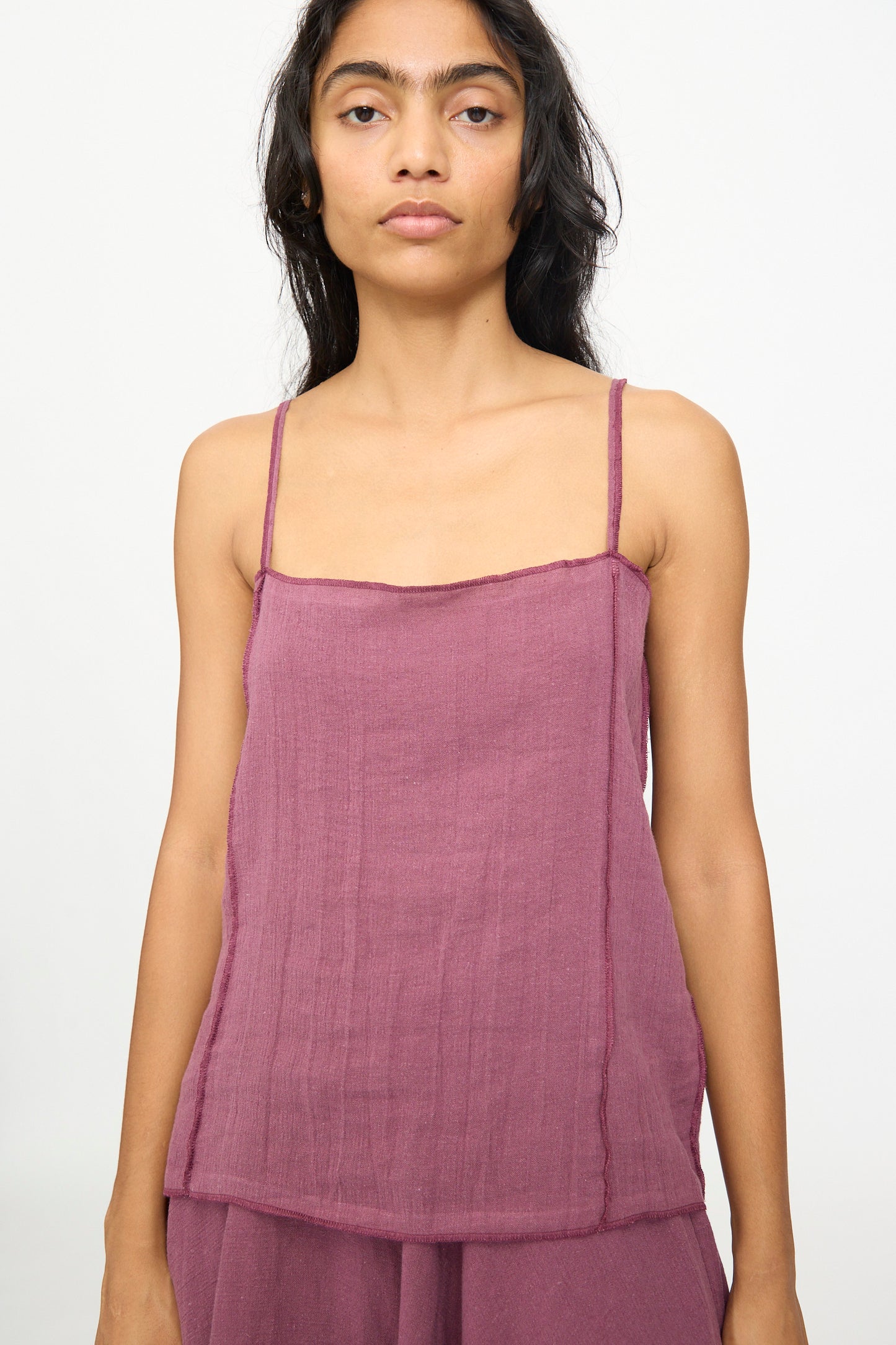 A person with long dark hair wears the Baserange Shok Slip Top in Aubergine, a relaxed fit tank with thin straps, paired with matching bottoms and stands against a plain light background.