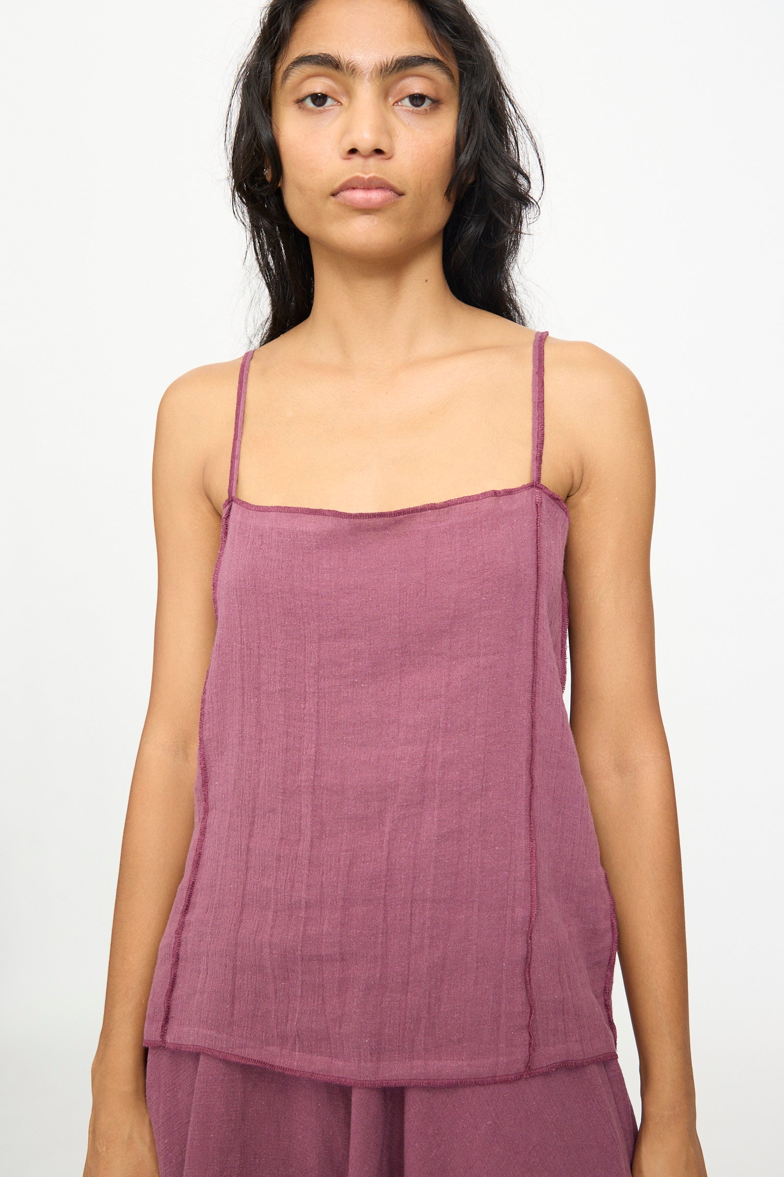 A person with long dark hair wears the Baserange Shok Slip Top in Aubergine, a relaxed fit tank with thin straps, paired with matching bottoms and stands against a plain light background.