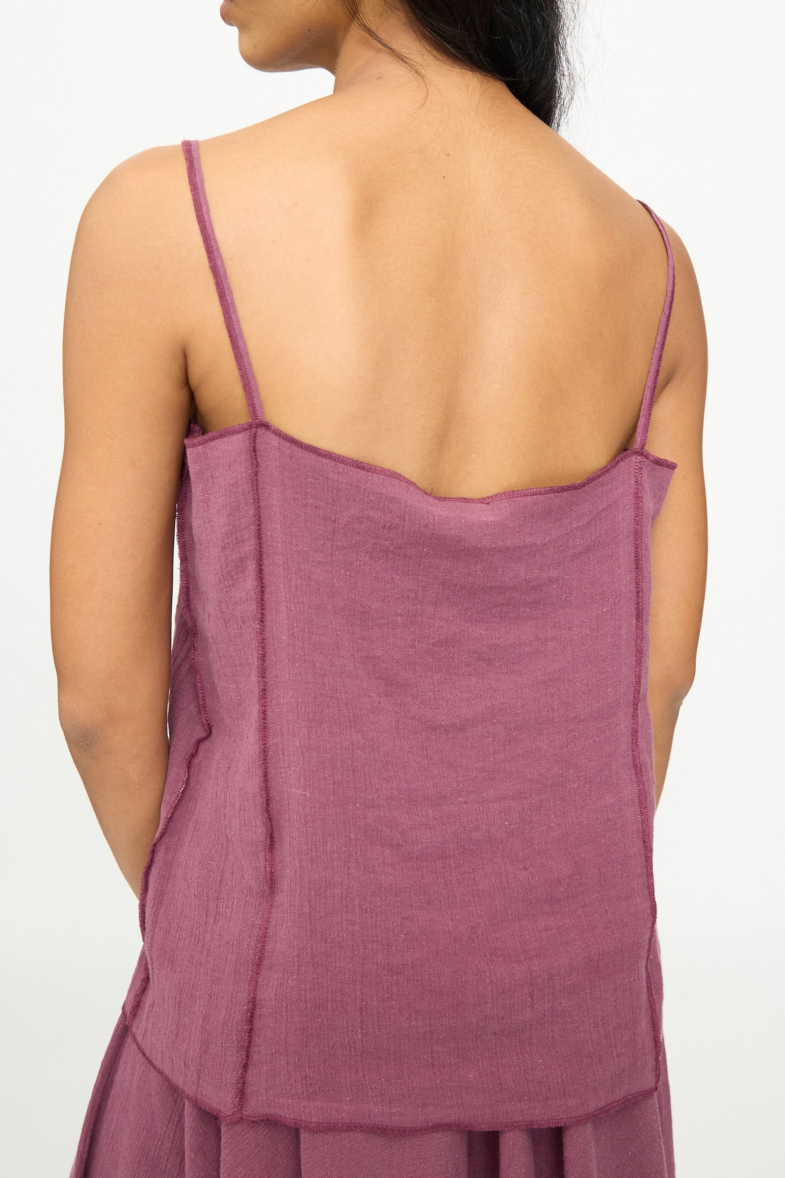 A person with long dark hair is photographed from behind against a plain background, wearing the Baserange Shok Slip Top in Aubergine—a relaxed fit tank with spaghetti straps.