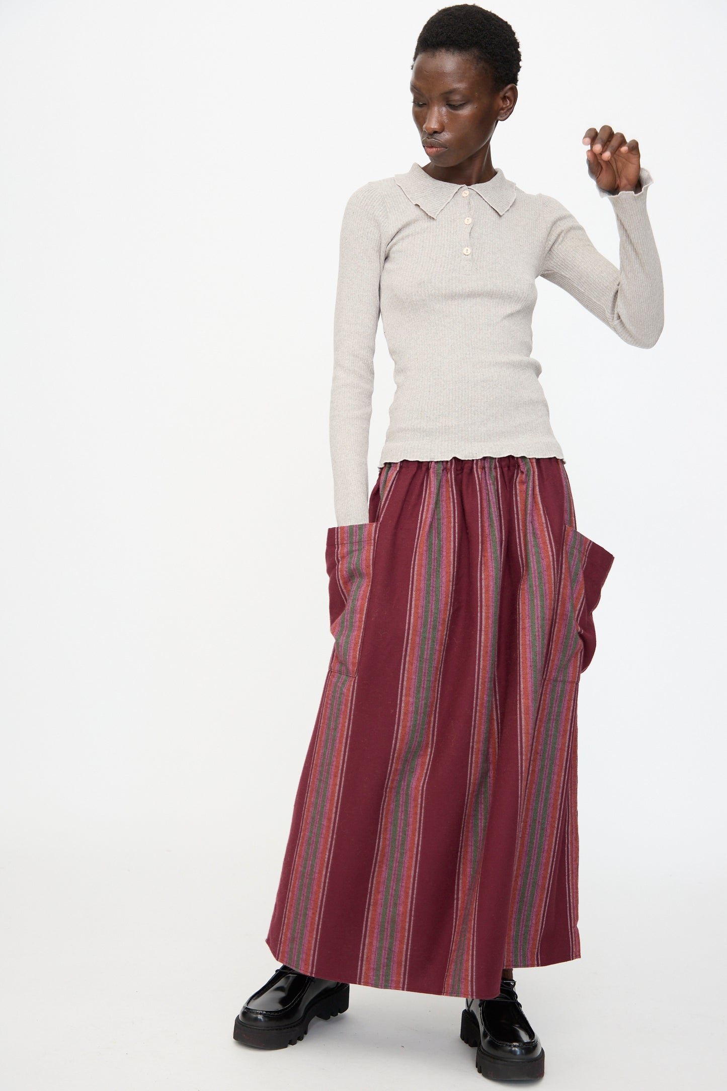A person wears the Baserange Snell Skirt in Maroon Stripe—an oversized, maroon striped cotton skirt with large pockets—paired with a light gray long-sleeve polo and black shoes, standing against a plain background.