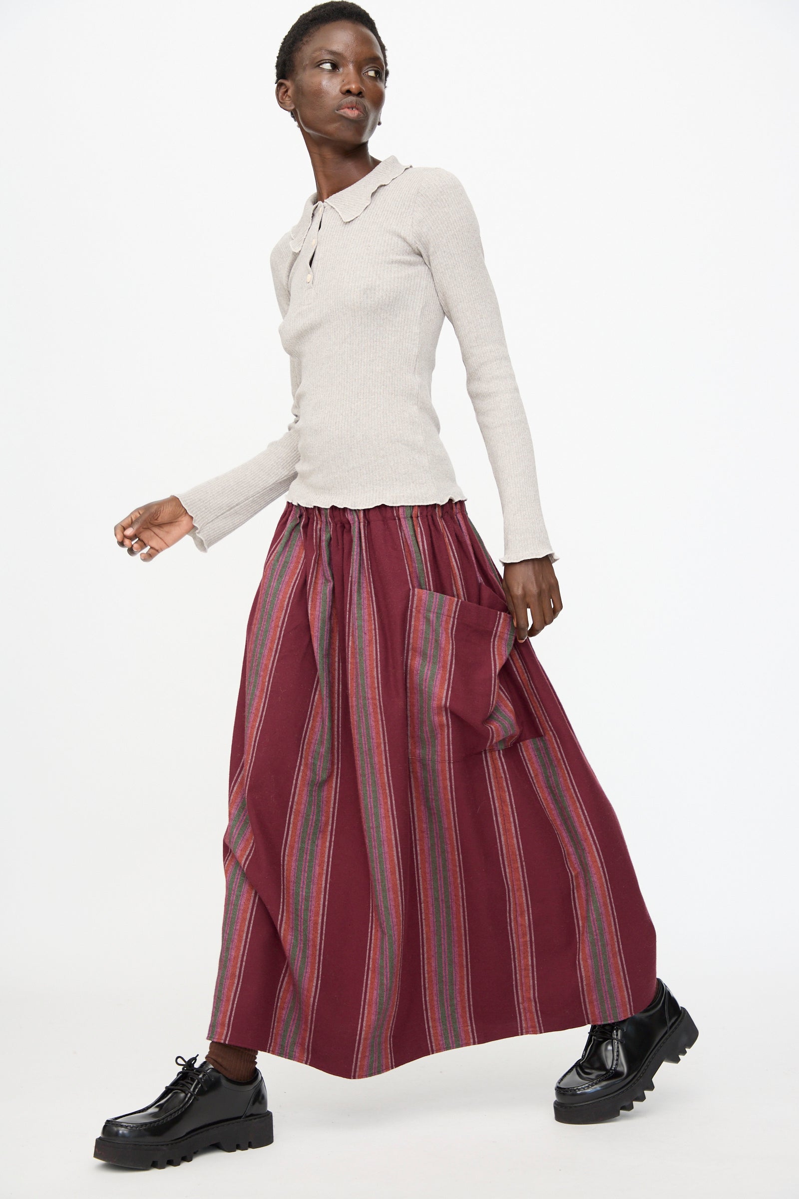 A person wearing a light long-sleeve top and the Baserange Snell Skirt in Maroon Stripe, featuring pockets, walking in black platform shoes against a plain white background.