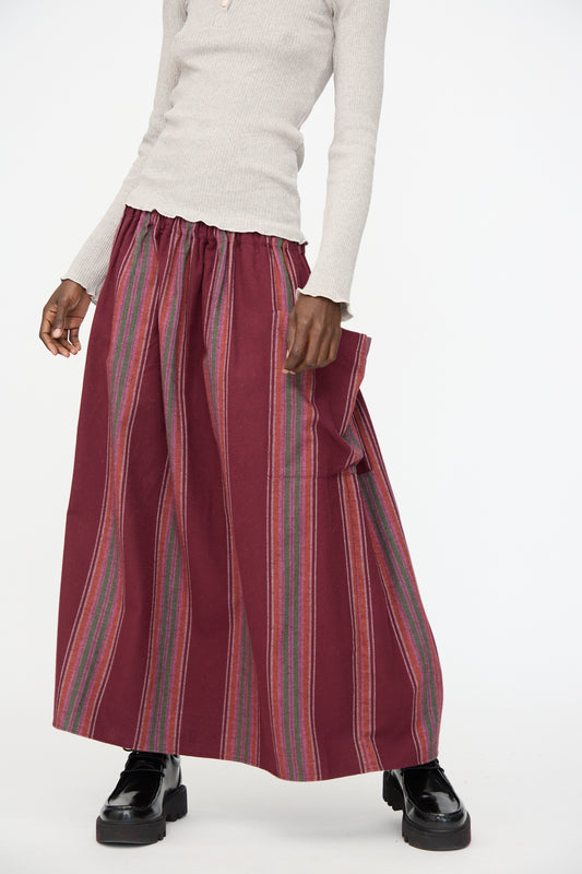 A person wearing a cream ribbed long-sleeve top, Baserange's Snell Skirt in Maroon Stripe with pockets, and black platform shoes stands against a plain light background.