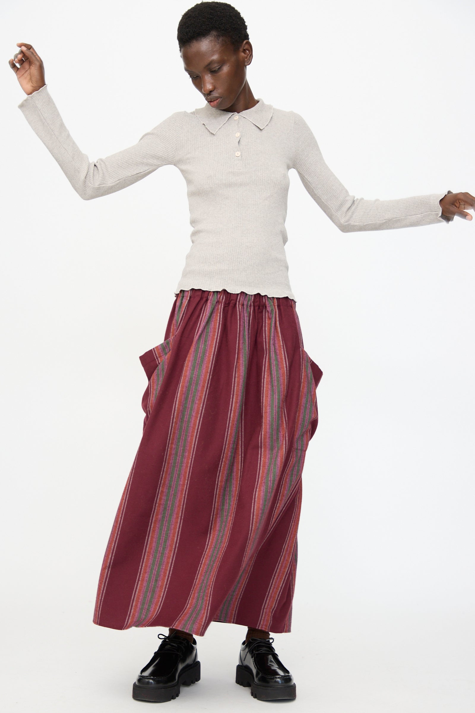 A person wearing a light gray long-sleeve shirt, black platform shoes, and the Baserange Snell Skirt in Maroon Stripe stands against a plain white background.