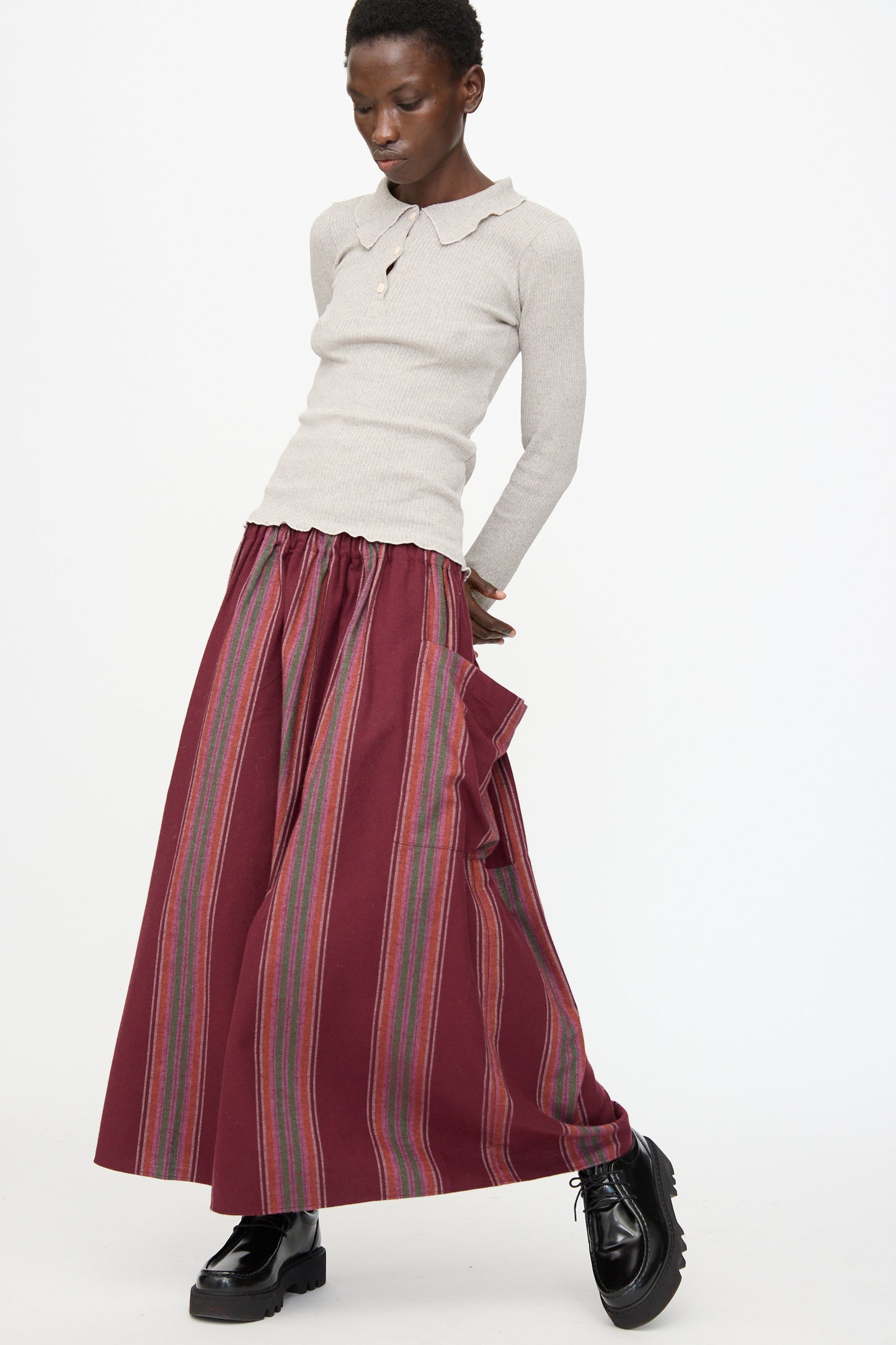 A person stands against a white background wearing a beige long-sleeve top, the Baserange Snell Skirt in Maroon Stripe with ruffle detail, and black platform shoes.