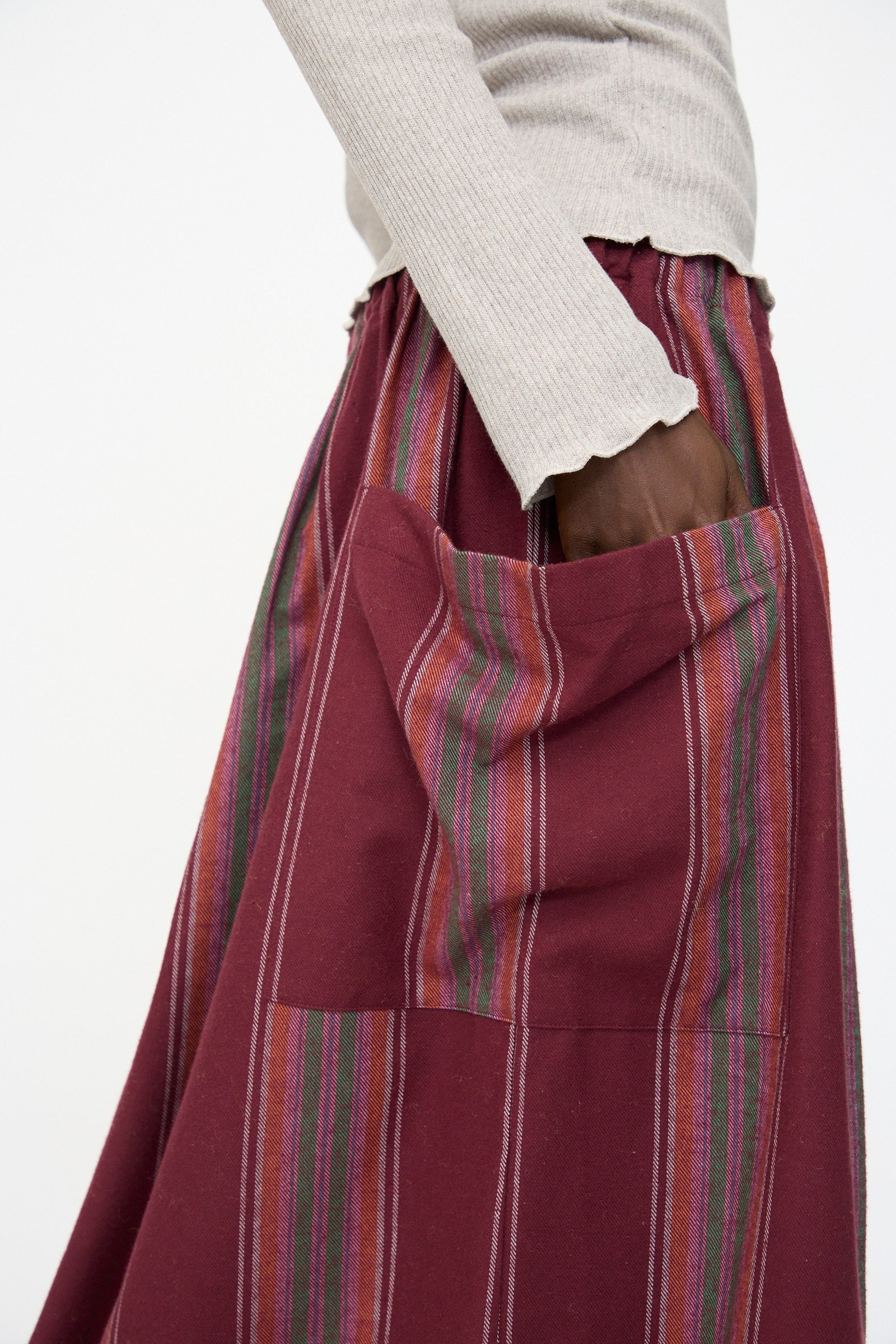 A person wearing a beige ribbed long-sleeve top and the Baserange Snell Skirt in Maroon Stripe, an oversized fit skirt with multicolored vertical stripes, with one hand in the pocket.