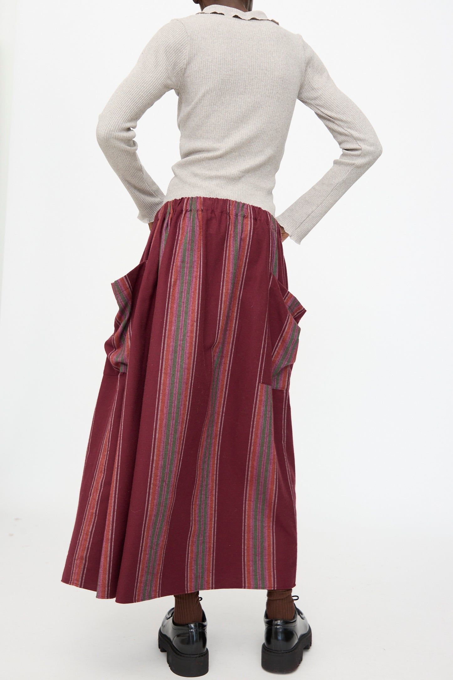 A person stands with hands on hips, shown from the back, wearing a light long-sleeve top and the Baserange Snell Skirt in Maroon Stripe, which features vertical stripes and large side pockets.