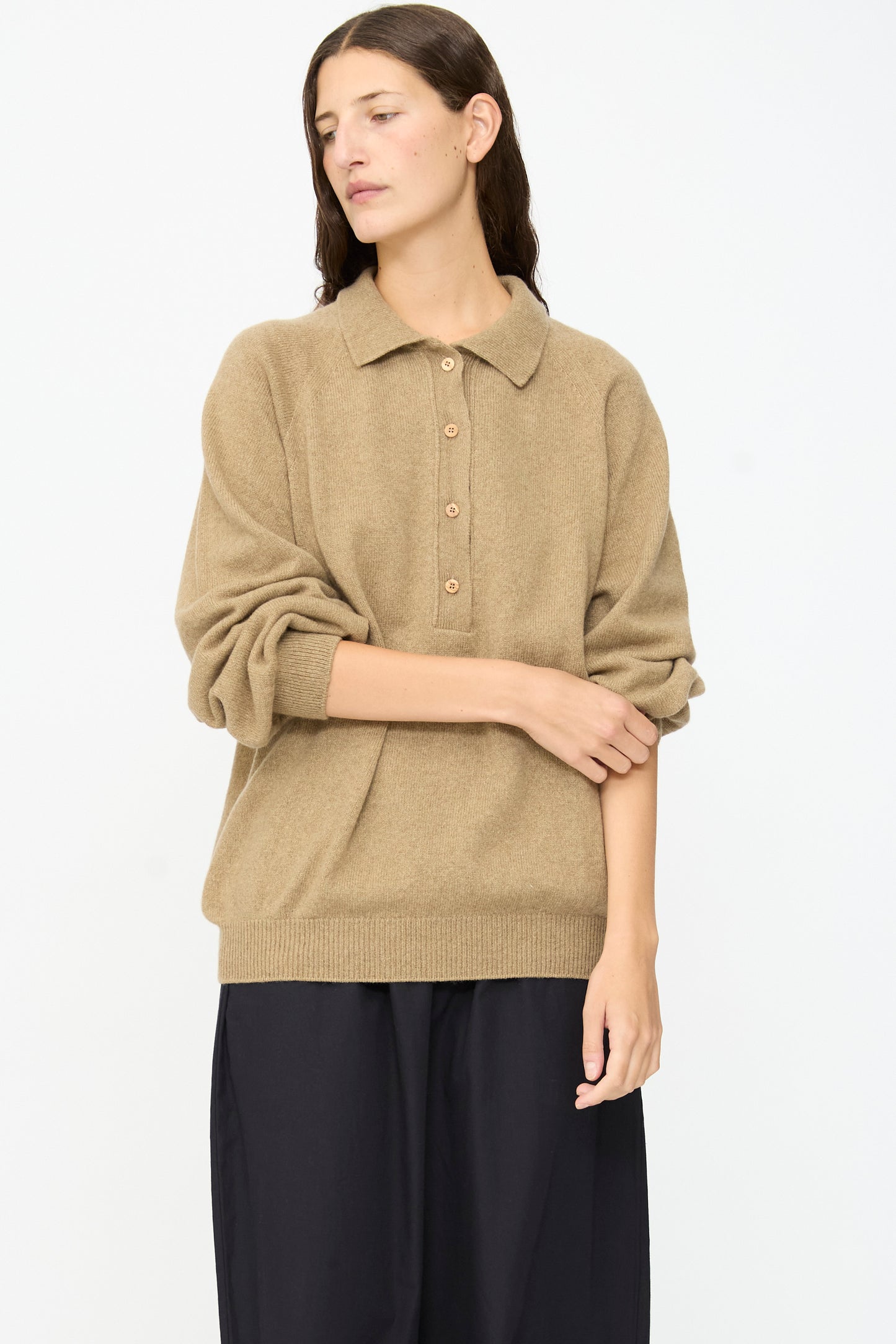 A person with long brown hair wears the Baserange Soar Polo in Rovere Brown, a tan cotton knit buttoned sweater with the sleeves pushed up, paired with a black skirt, standing against a plain white background.