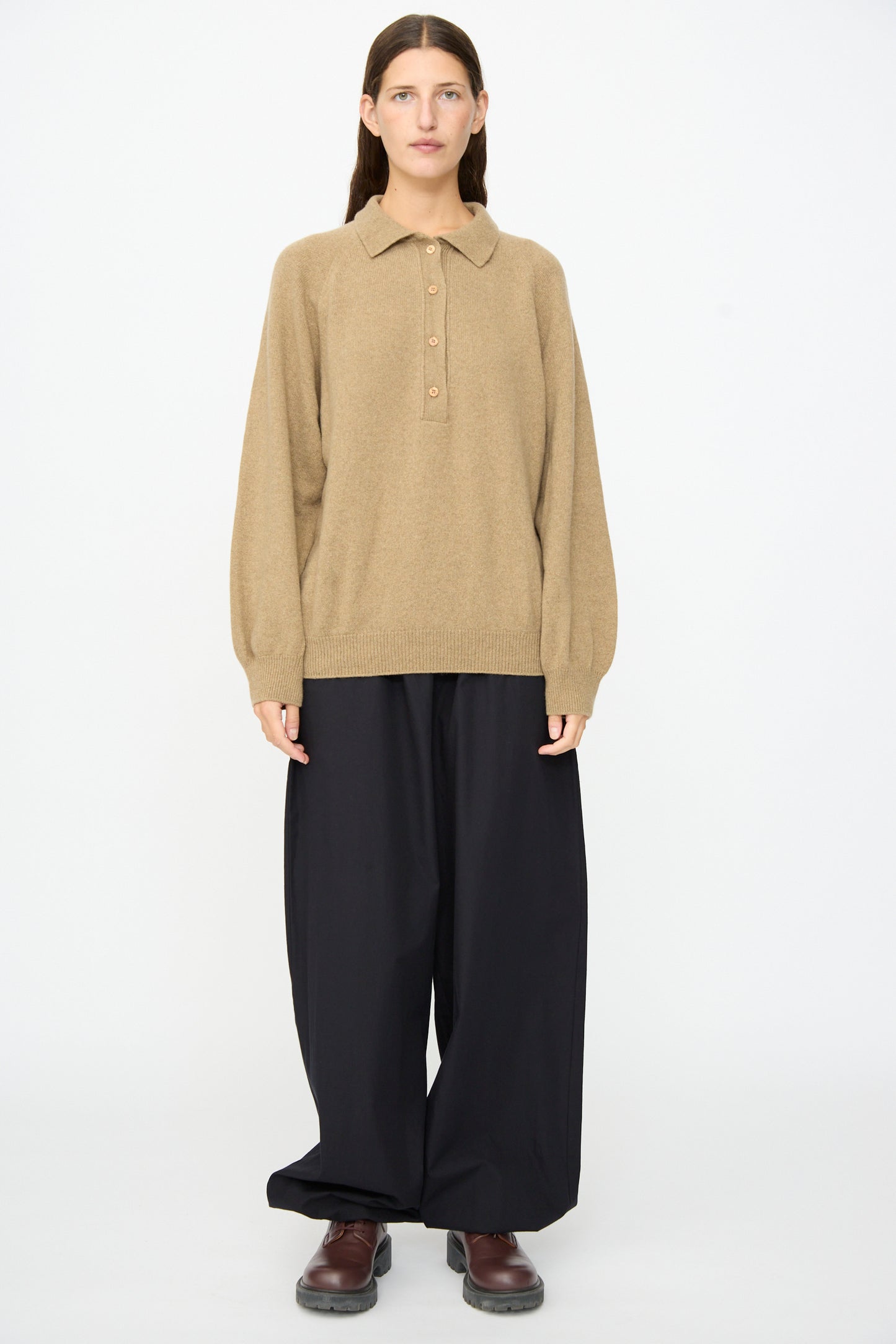 A person stands against a plain background, wearing the Soar Polo in Rovere Brown by Baserange, paired with wide black trousers and brown shoes, with their hands at their sides and a neutral expression.