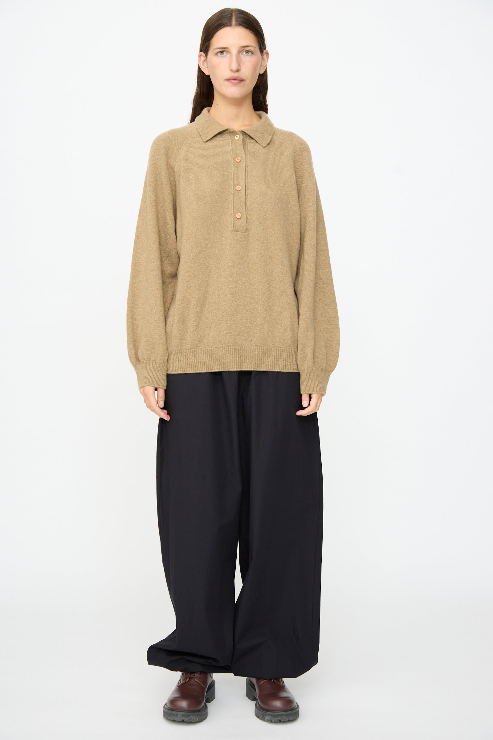A person stands against a plain background, wearing the Soar Polo in Rovere Brown by Baserange, paired with wide black trousers and brown shoes, with their hands at their sides and a neutral expression.